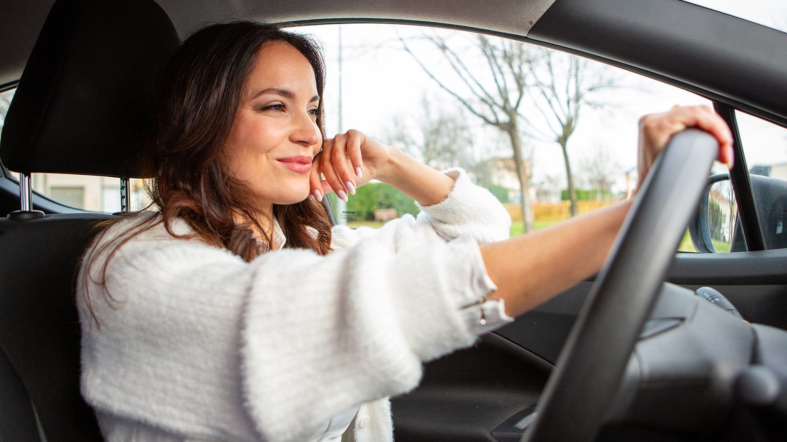 A woman with confident personality traits sits in the driver’s seat of a car, smiling and looking out the window, one hand driving. She wears a white sweater, with trees and houses visible outside.