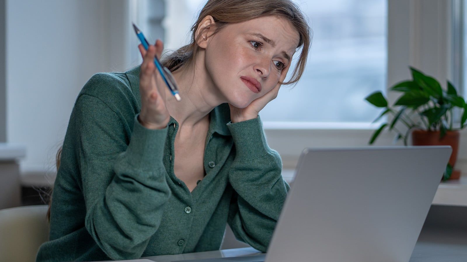 A woman sits at a desk in front of a laptop, looking confused and frustrated, with her head resting on one hand and holding a pen in the other. A plant is on the windowsill in the background.