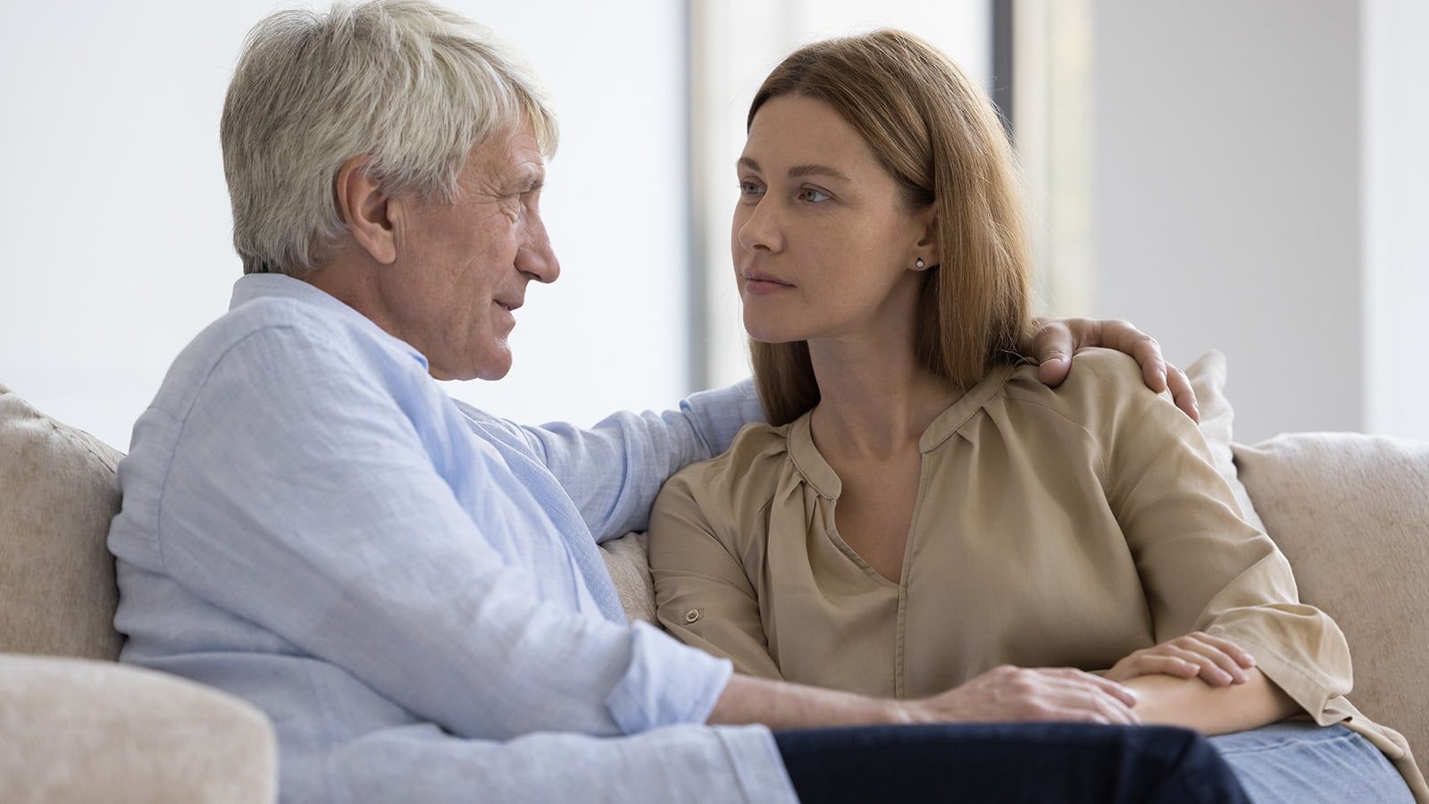 An older boomer father and a younger woman sit close together on a couch, facing each other and having a serious conversation about rent. The man’s arm is around the woman’s shoulders, and they both appear thoughtful.