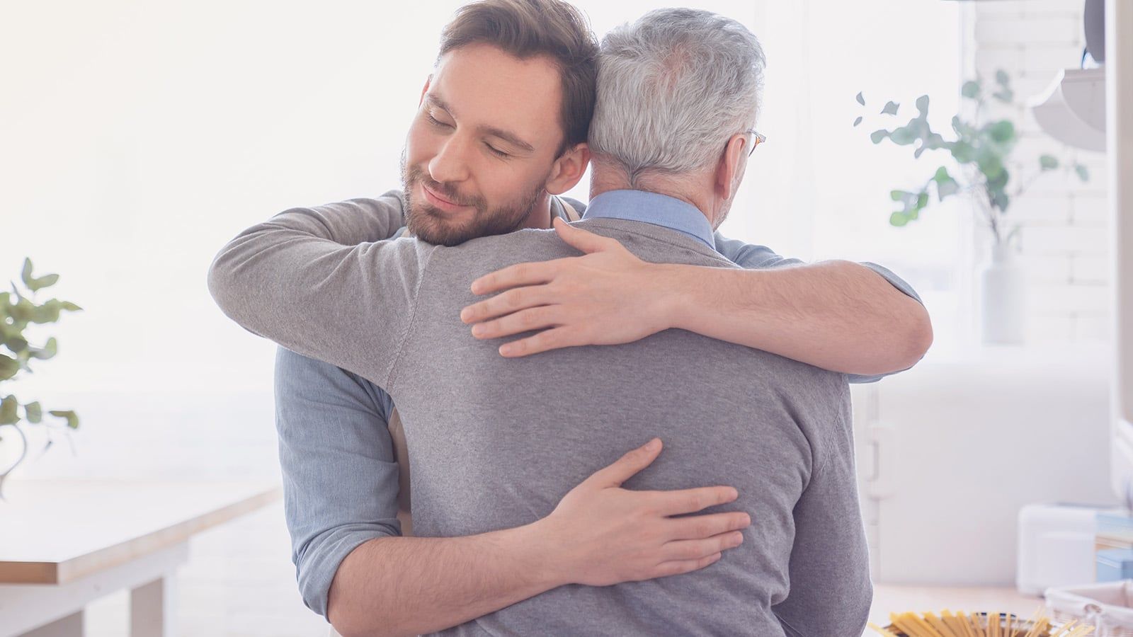 A younger man warmly hugs an older man with gray hair in a bright, cozy kitchen. Both appear content and comfortable, sharing a moment that beautifully reflects the bond of parenthood amid soft natural light and plants in the background.