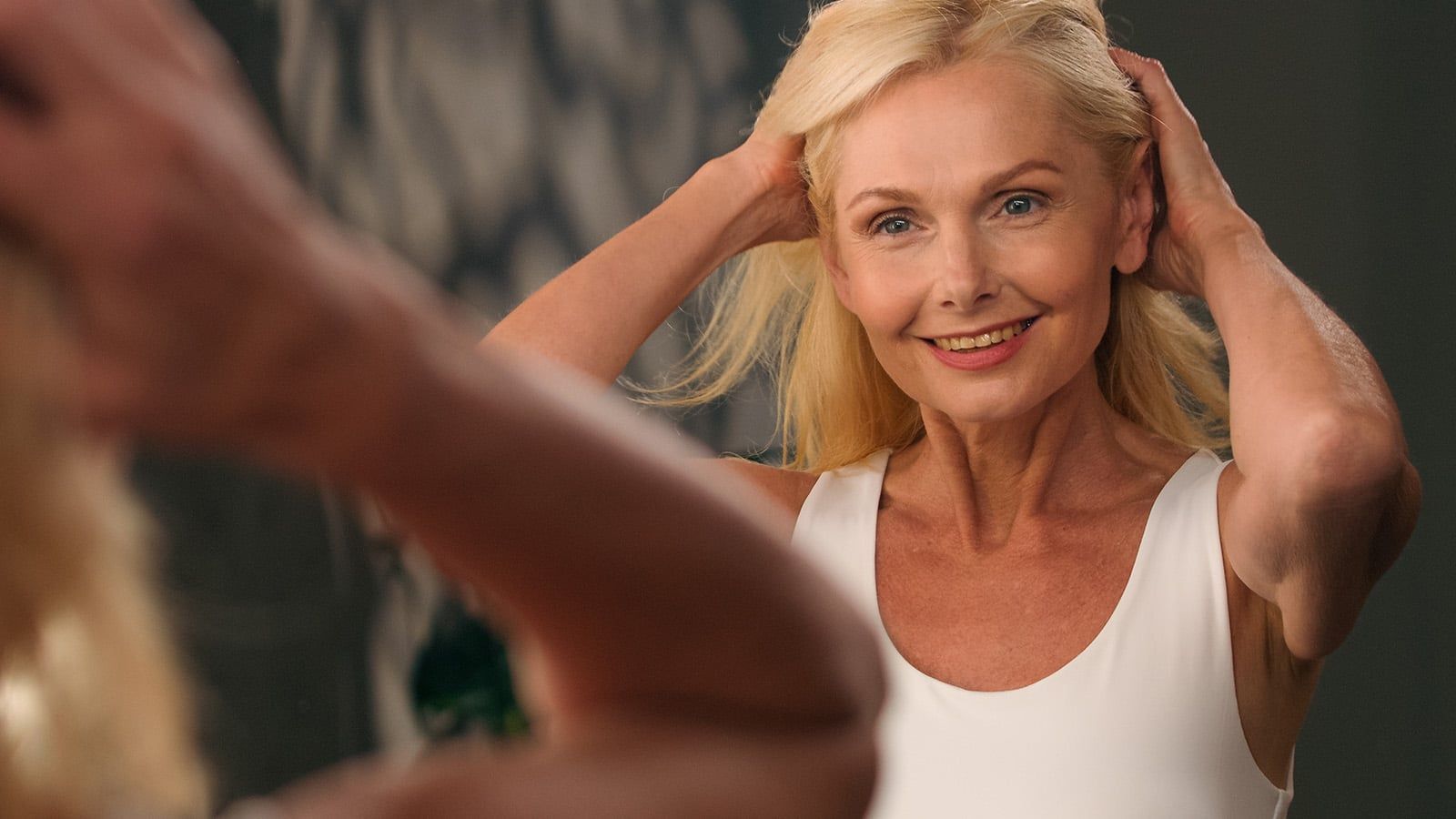 A smiling woman in her 60s with blonde hair stands in front of a mirror, touching her hair with both hands and wearing a white tank top. Her reflection captures the quiet confidence and invisible expertise that come with aging gracefully.