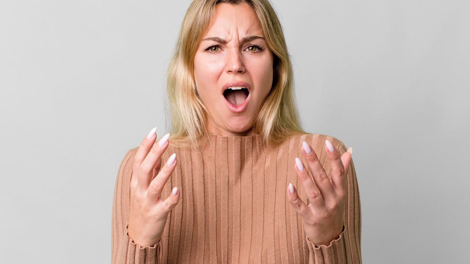 A woman with long blonde hair wearing a beige ribbed sweater looks surprised or upset, as if she's setting boundaries or saying no, her mouth open and hands raised in front of her against a plain light gray background.