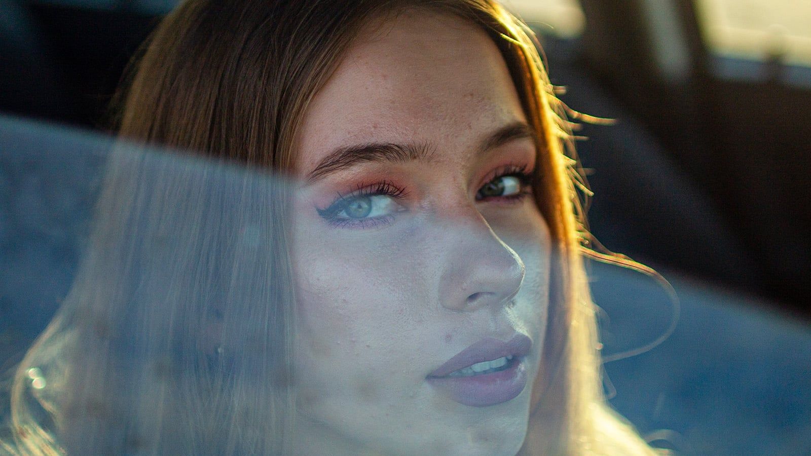 A close-up of a young woman with long brown hair and blue eyes, looking through a car window as sunlight softly illuminates her face; the hazy, reflective effect hints at overcoming difficulty, capturing quiet resilience.