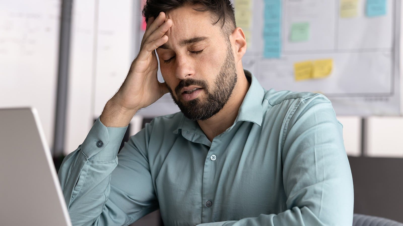 A man with a beard in a teal shirt sits at a desk, appearing stressed and employee overworked, with his eyes closed and hand on his forehead, as his laptop and whiteboard sit in the background.