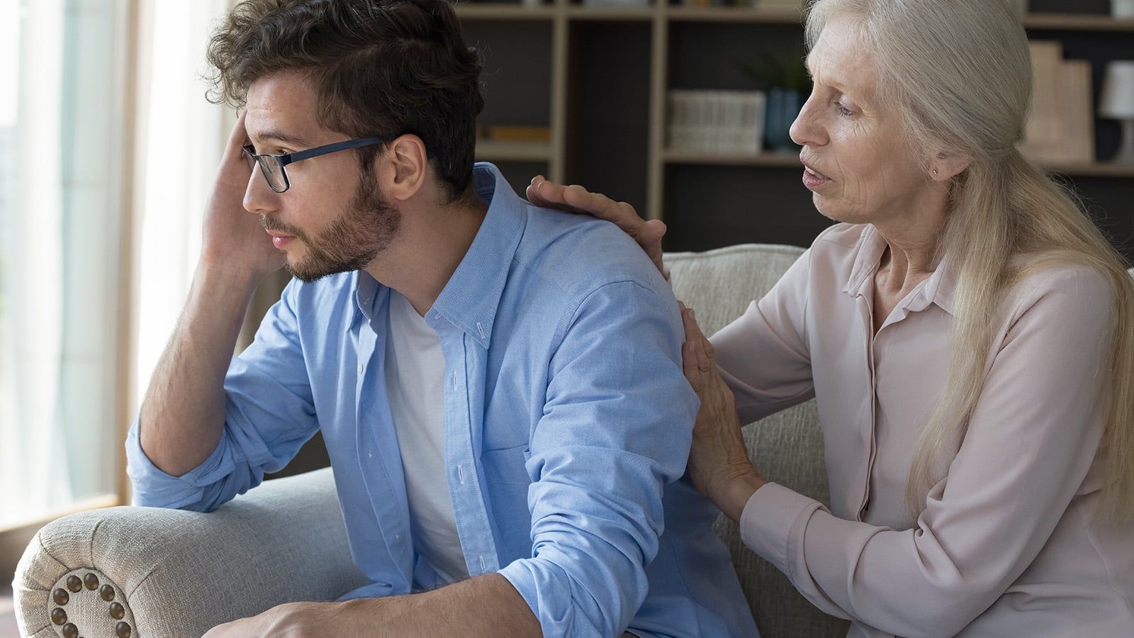 An older woman, likely his parent, sits beside a younger man on a couch, gently touching his shoulder and back as he looks stressed. Their serious conversation hints at navigating toxic patterns in adulthood.