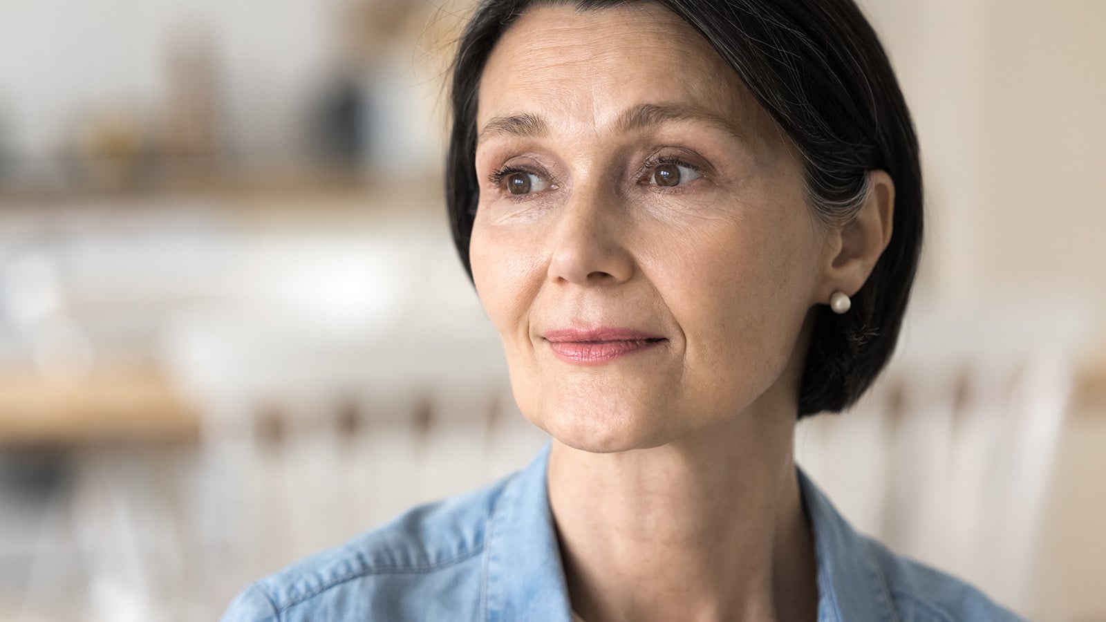 A middle-aged woman with short dark hair and pearl earrings looks thoughtfully into the distance, perhaps reflecting on generational parenting, wearing a light blue shirt with a softly blurred indoor background.