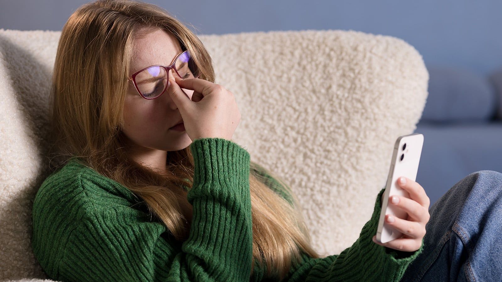 A young woman wearing glasses and a green sweater sits on a couch, holding a smartphone in one hand and pinching the bridge of her nose with her other hand, showing signs of exhaustion that often affect the nervous system.