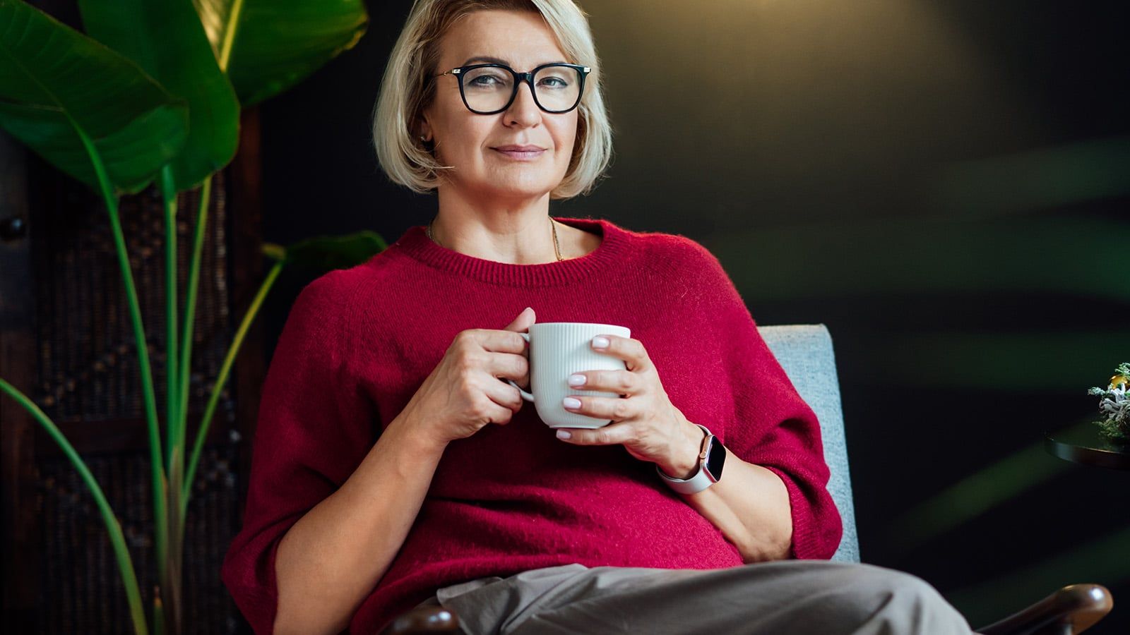 A woman with short blonde hair and glasses, wearing a red sweater and beige pants, sits in a chair holding a white mug, looking at the camera—her calm gaze reflecting her life story of being alone. Green plants and a dark wall are in the background.