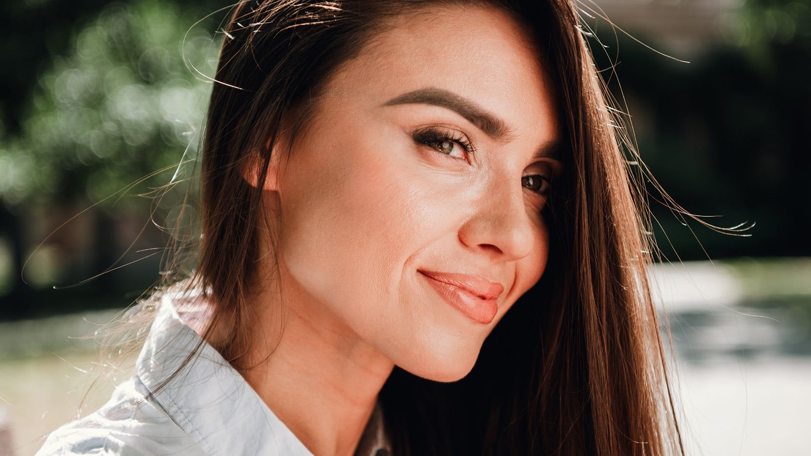 A woman with long brown hair and natural makeup smiles softly at the camera outdoors, surrounded by greenery and sunlight, radiating calm as she enjoys a peaceful moment of coping with burnout.