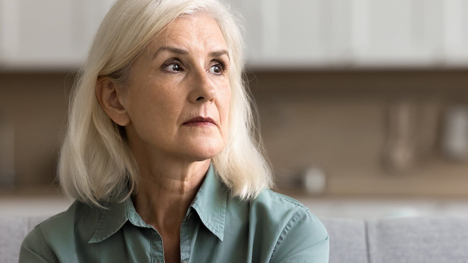 An older woman with white hair and a light green shirt sits indoors, looking thoughtfully to the side with a serious expression—capturing a quiet moment of emotional openness. The background is softly blurred.