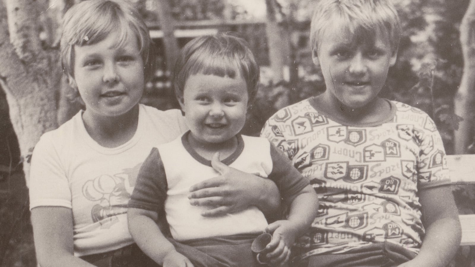 Three children sit closely together on a bench outdoors. The child in the middle, a toddler, smiles while being held by the older children on either side—a charming black and white image reflecting generational differences.