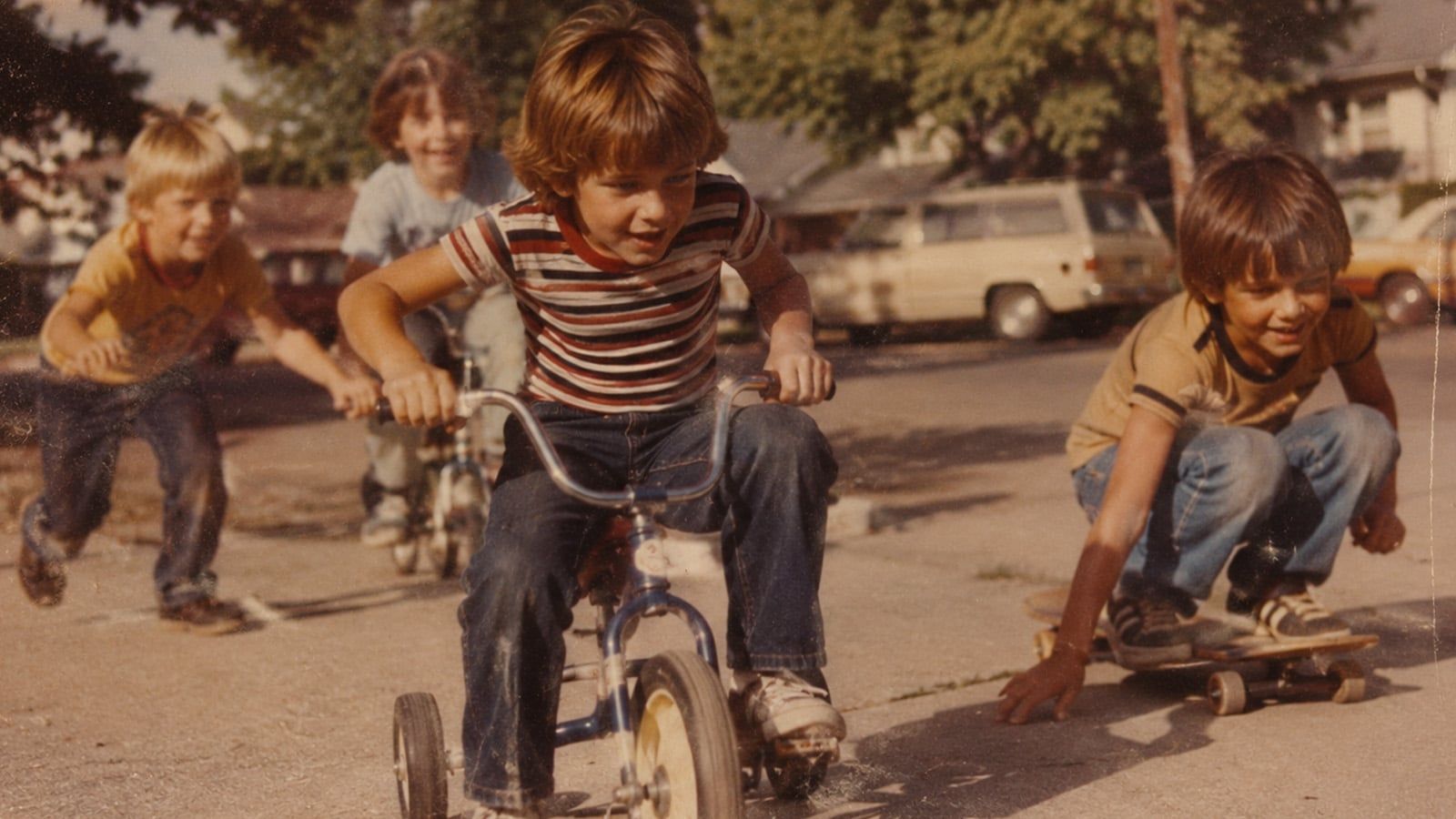 Four children play outside; one rides a tricycle in front, another is on a skateboard, and two run behind. Parked cars and trees set the scene, evoking life lessons and nostalgic memories of the 70s and 80s.