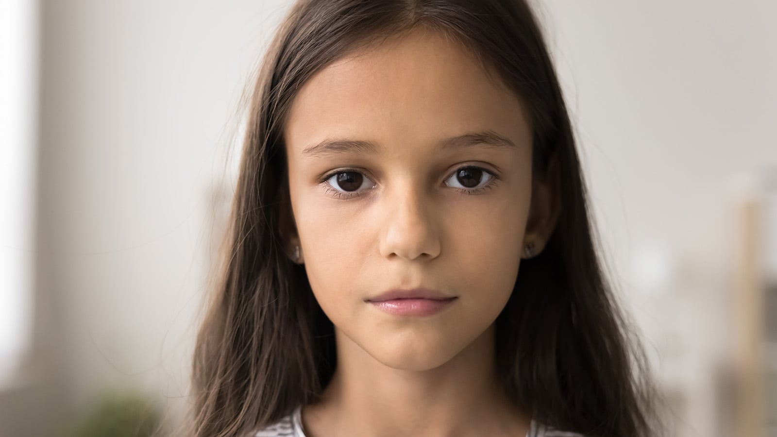 A young girl with long brown hair and brown eyes, shaped by quiet traits, looks directly at the camera with a neutral expression. She is indoors, with a softly blurred background hinting at early brushes with adulthood.