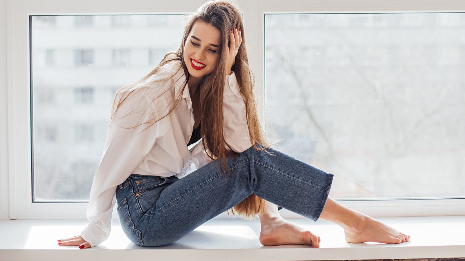 A woman with long brown hair, wearing a white shirt and blue jeans, sits barefoot on a windowsill, smiling and gently touching her head—lost in solitude as soft natural light streams through the window behind her.