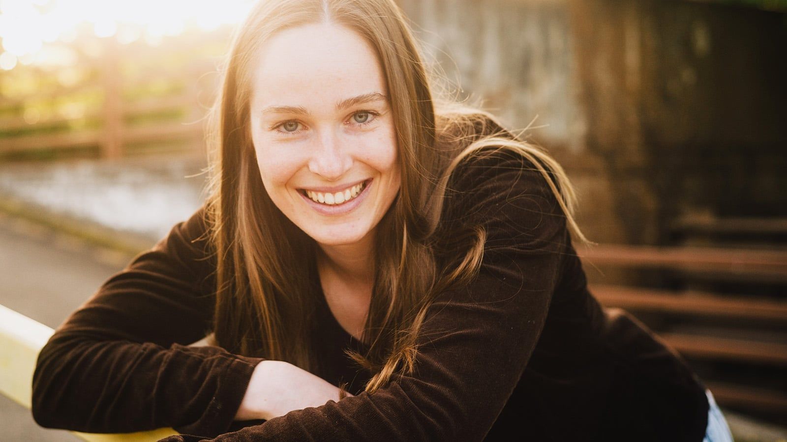 A young woman with long brown hair and a black top smiles warmly at the camera outdoors, sunlight shining in the background, radiating an air of lovability.