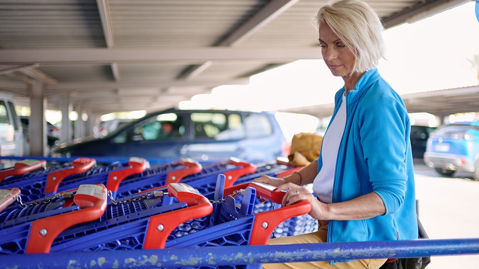 A woman with short blonde hair stands under a parking structure, retrieving a shopping cart from a row of blue carts with red handles, her focused expression hinting at character traits of determination and organization. Several cars are parked in the background.