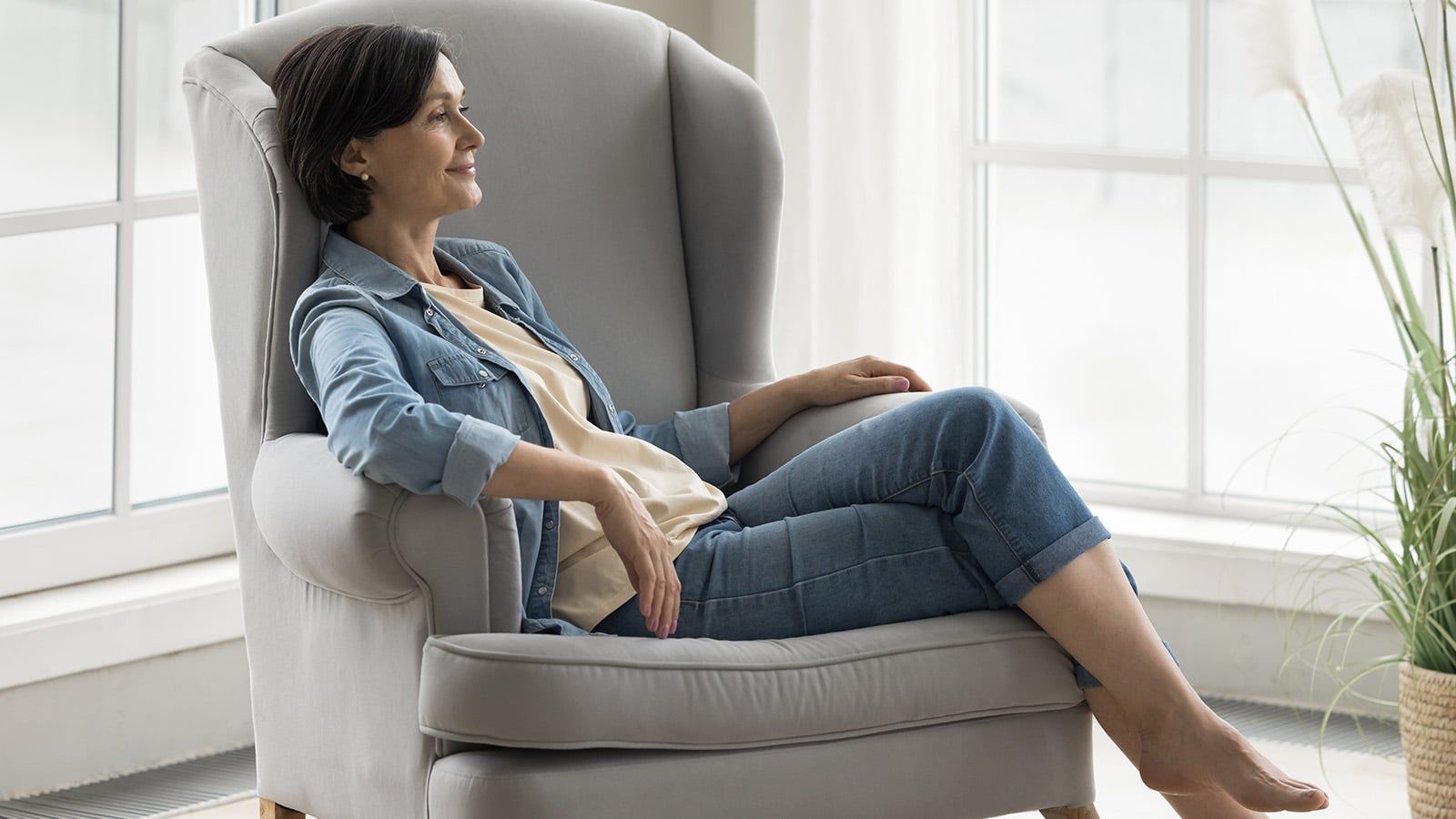 A woman with short dark hair relaxes in a gray armchair by large windows, wearing a denim jacket and jeans, smiling as she looks outside, enjoying the quiet phone and the comfort of a small circle after reaching your 60s.