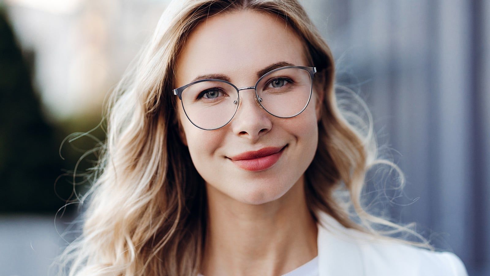 A woman with long blonde hair and glasses, displaying signs of intelligence, smiles softly at the camera in a white top while standing outdoors with a blurred background.