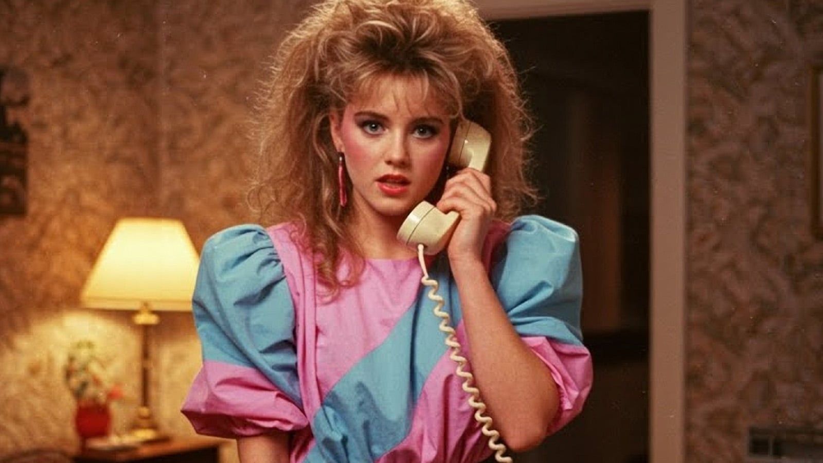 A woman with voluminous 1980s-style blonde hair and bold makeup wears a pink and blue puff-sleeve dress, holding a kitchen phone to her ear in a retro-decorated room with a lamp and patterned wallpaper.
