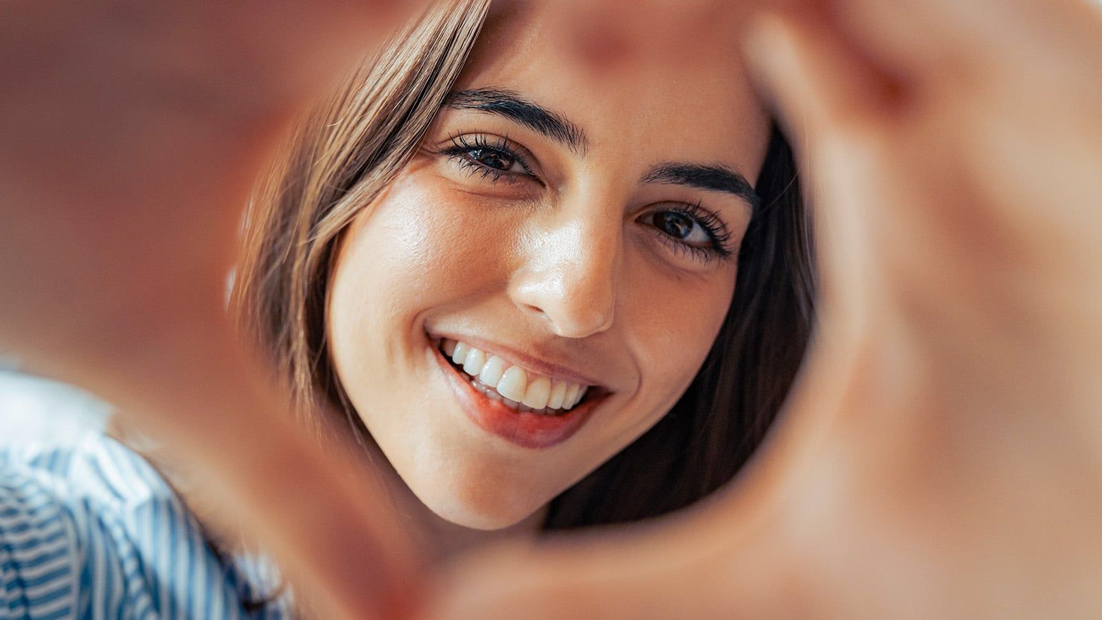 A young woman with long brown hair smiles warmly at the camera, her hands forming a heart shape in the foreground, framing her face. She appears happy and relaxed in a striped shirt, radiating the joy that comes from close relationships and human happiness.