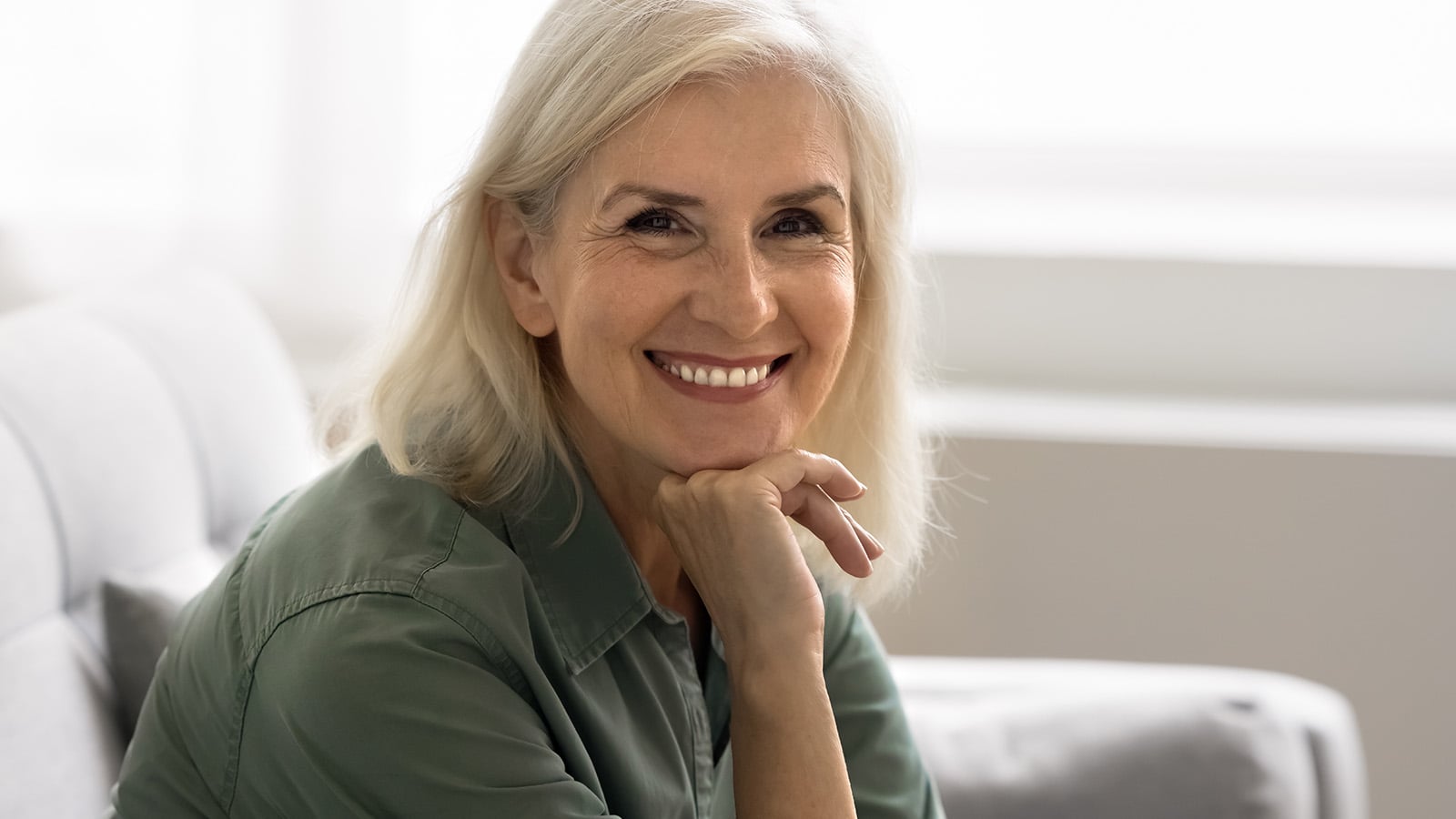 A smiling older woman with gray hair, embodying healthy aging, sits on a light-colored sofa, resting her chin on her hand. She wears a green shirt and looks directly at the camera. The background is softly lit and out of focus.