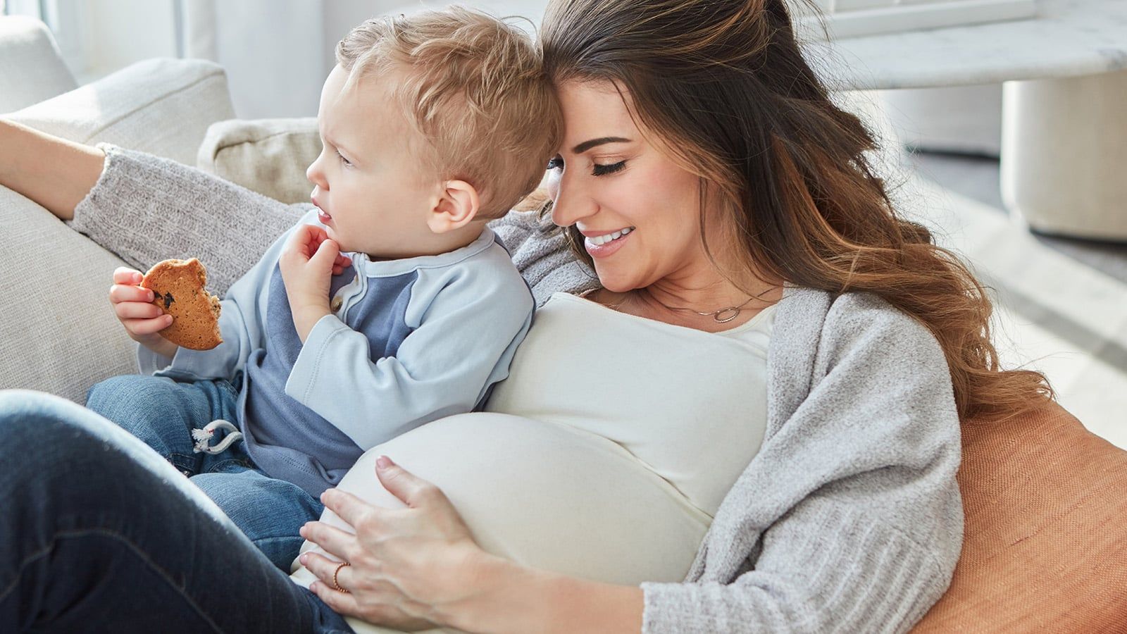A pregnant woman smiles while relaxing on a couch with a young child who is eating a cookie. They both appear happy and comfortable, sharing a joyful moment that beautifully contrasts common feelings of parenthood guilt.