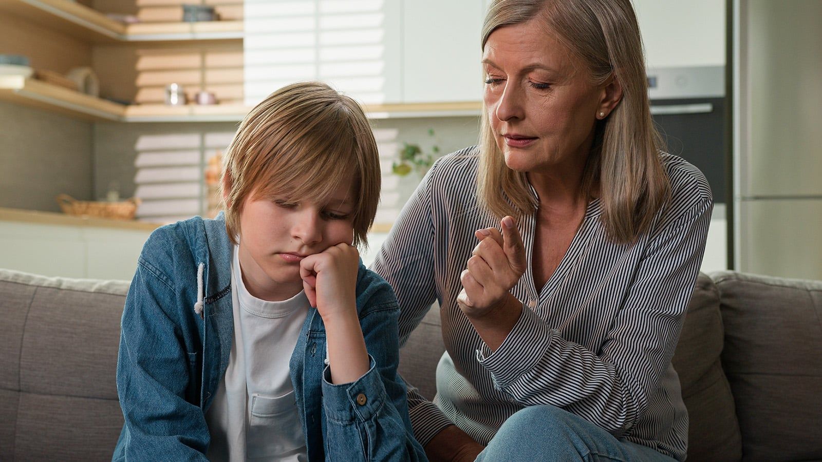A concerned aging parent sits on a couch beside a sad or worried young boy, gently talking to him and offering comfort in a bright, modern living room, reflecting on changing family patterns.