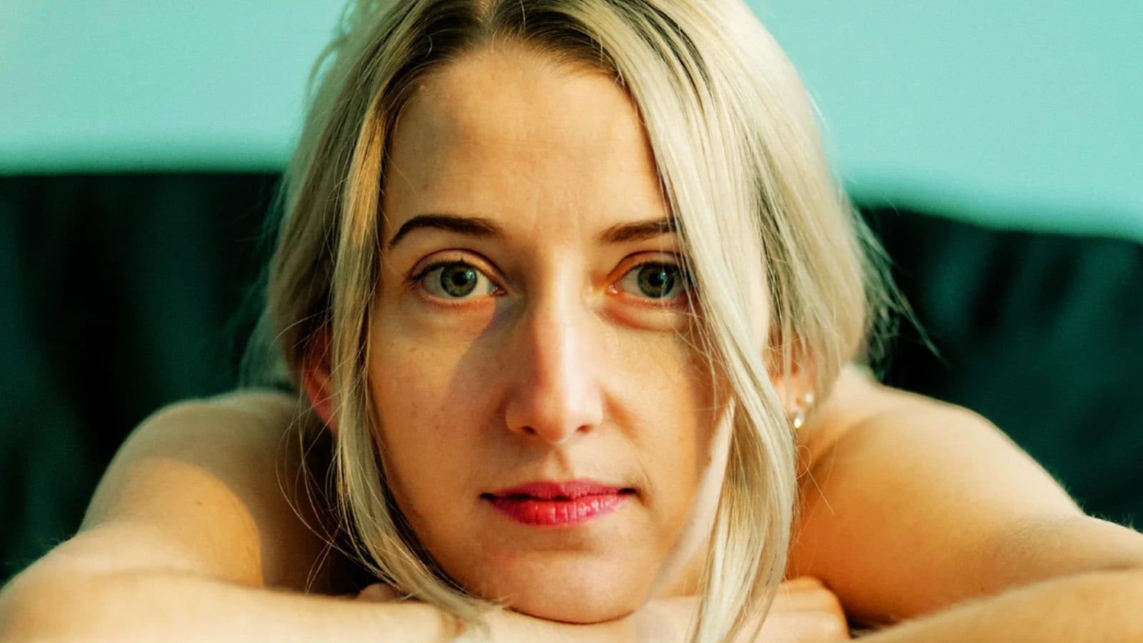 A woman with blonde hair rests her chin on her hands, gazing forward with a thoughtful expression that hints at loneliness. She is indoors, with a soft blue background and natural lighting on her face.