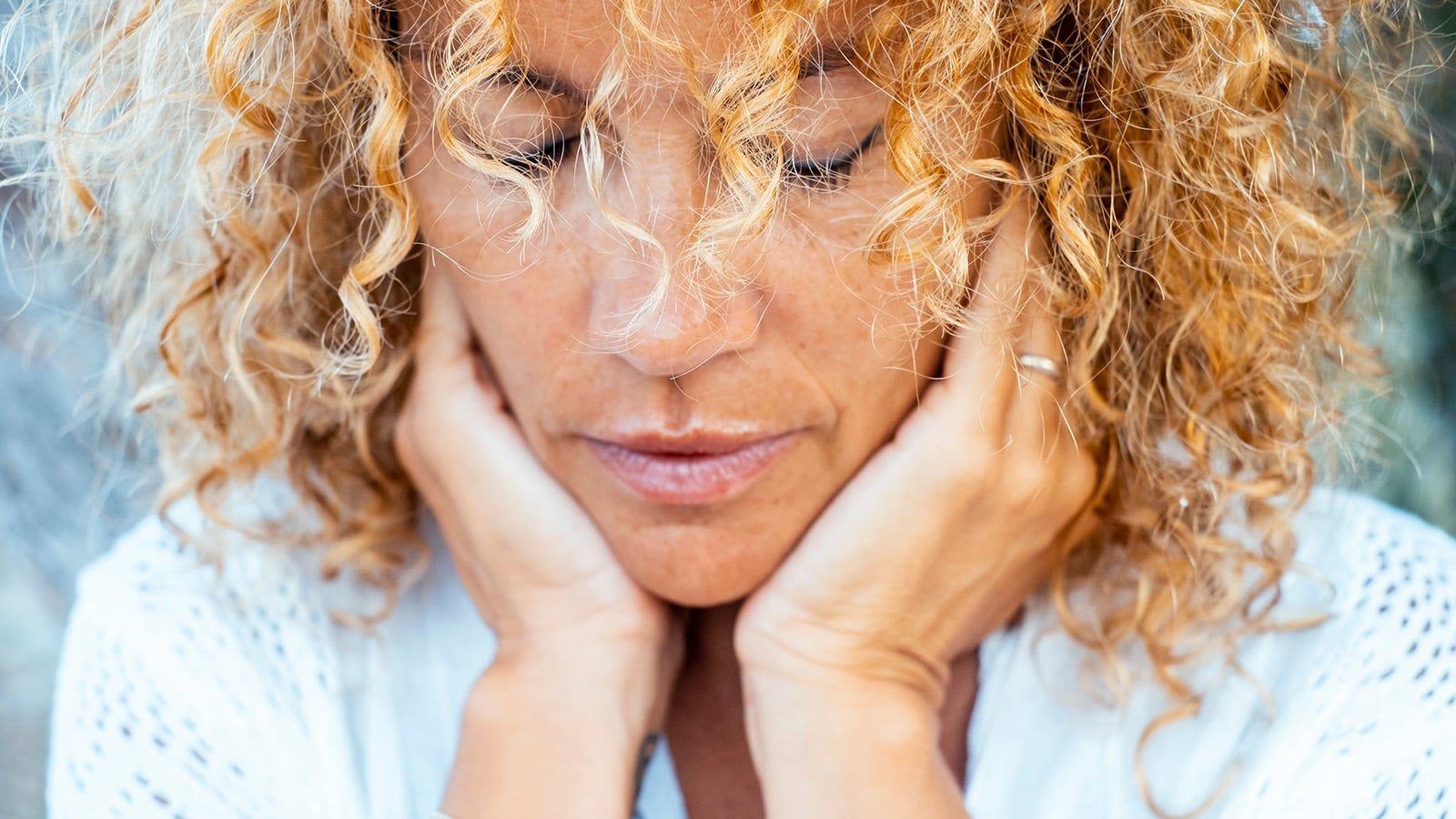 Close-up of a woman with curly blond hair resting her face in her hands, eyes closed, appearing thoughtful—perhaps feeling disconnected or reflecting on outgrowing relationships. She wears a white top and a ring on her left hand.