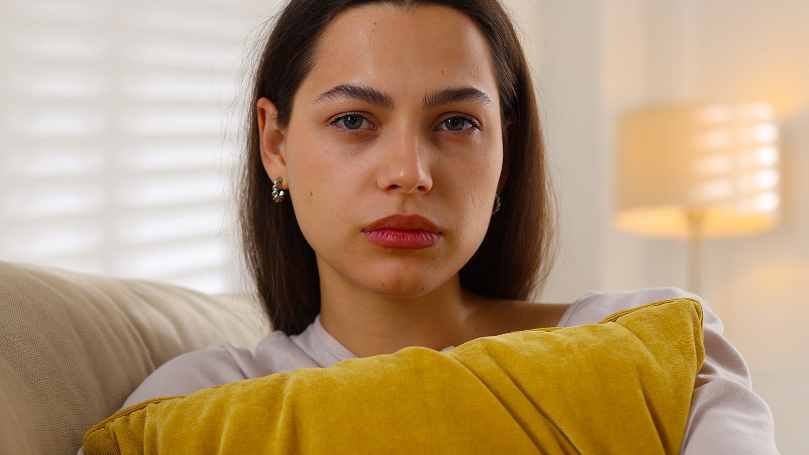 A woman with long brown hair, the eldest daughter, sits on a couch holding a yellow pillow. She has a neutral, slightly serious expression. A lamp and white blinds are visible in the softly lit background.