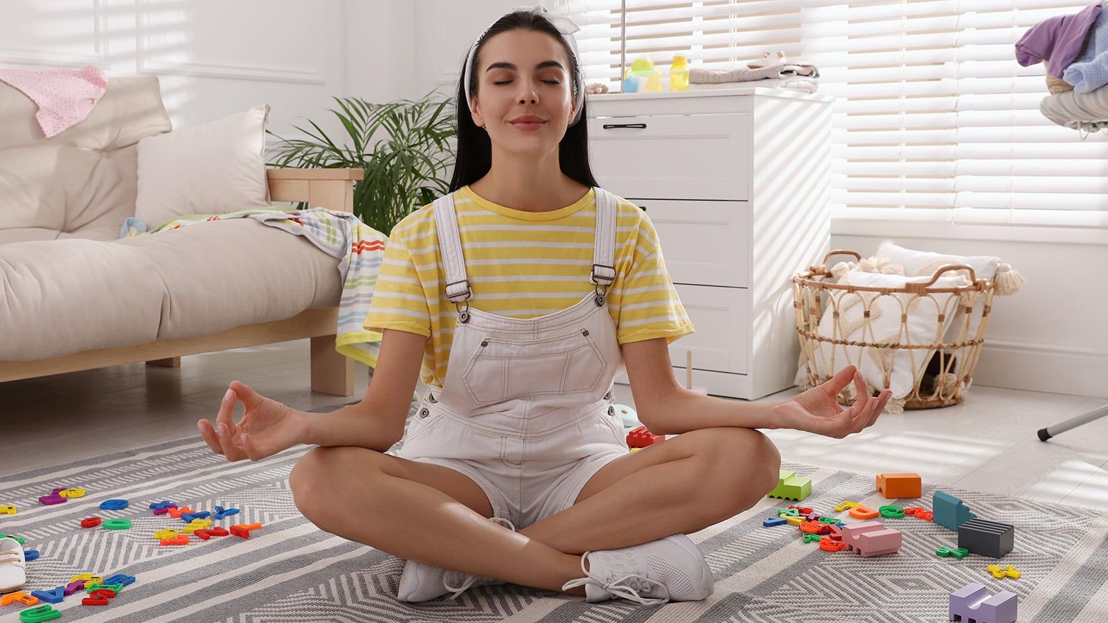 A woman in a striped yellow shirt and white overalls sits cross-legged on a rug, meditating with eyes closed, surrounded by colorful plastic toys in a bright living room that reflects real family dynamics and a happily lived-in home.