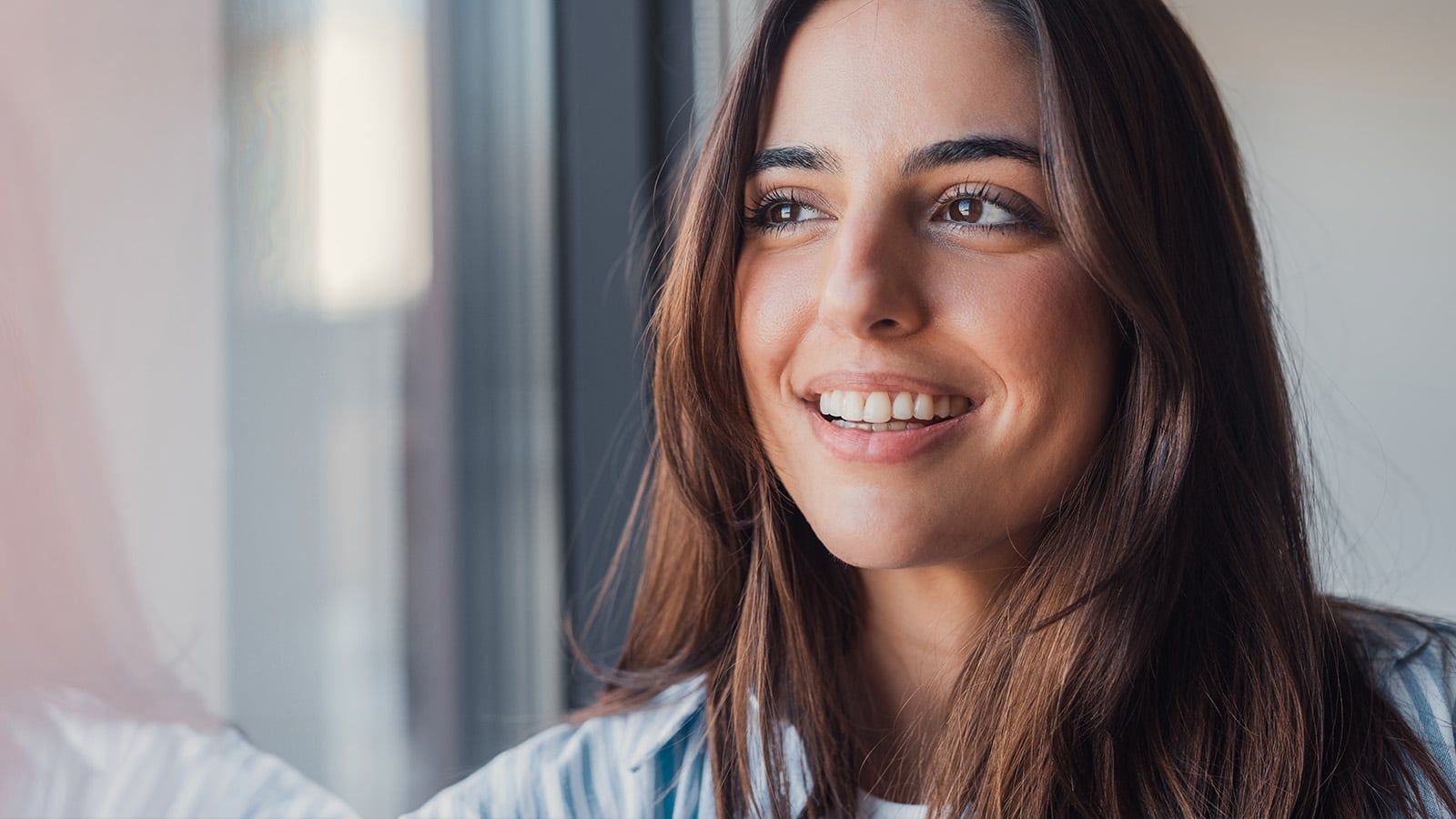 A young woman with long brown hair smiles while looking out of a window. She is wearing a light blue and white shirt, and natural light brightens her face, reflecting a hopeful expression amid emotional chaos.