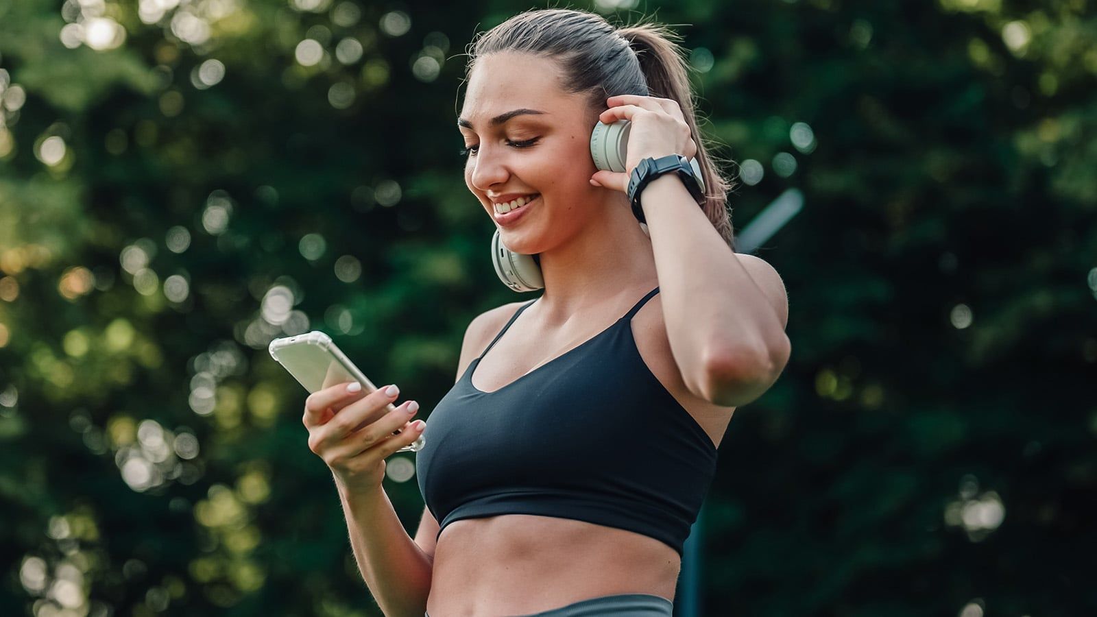 A smiling woman in athletic wear holds a smartphone and adjusts her headphones outdoors, embracing exercise amidst trees and sunlight.