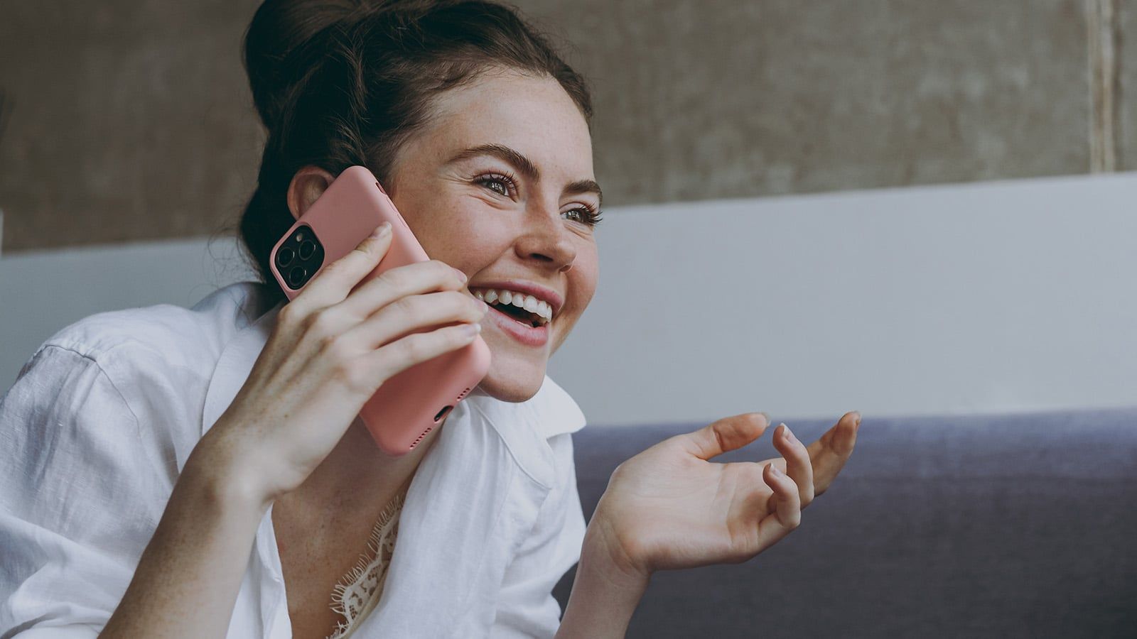 A smiling woman in a white shirt talks on her phone, holding it to her ear with one hand while gesturing with the other, sitting indoors on a sofa. Her warm expression suggests she's nurturing a close relationship, always being kind and attentive during conversations.