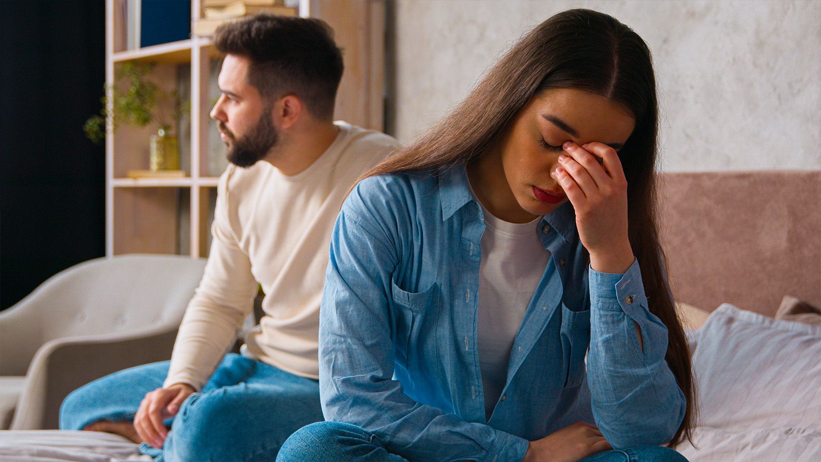 A woman sits on a bed, looking upset with her hand on her face, while a man sits behind her, turned away and gazing into the distance. Both appear distressed, suggesting a tense or emotional moment that may reflect challenges in personal growth or difficulties related to social habits.