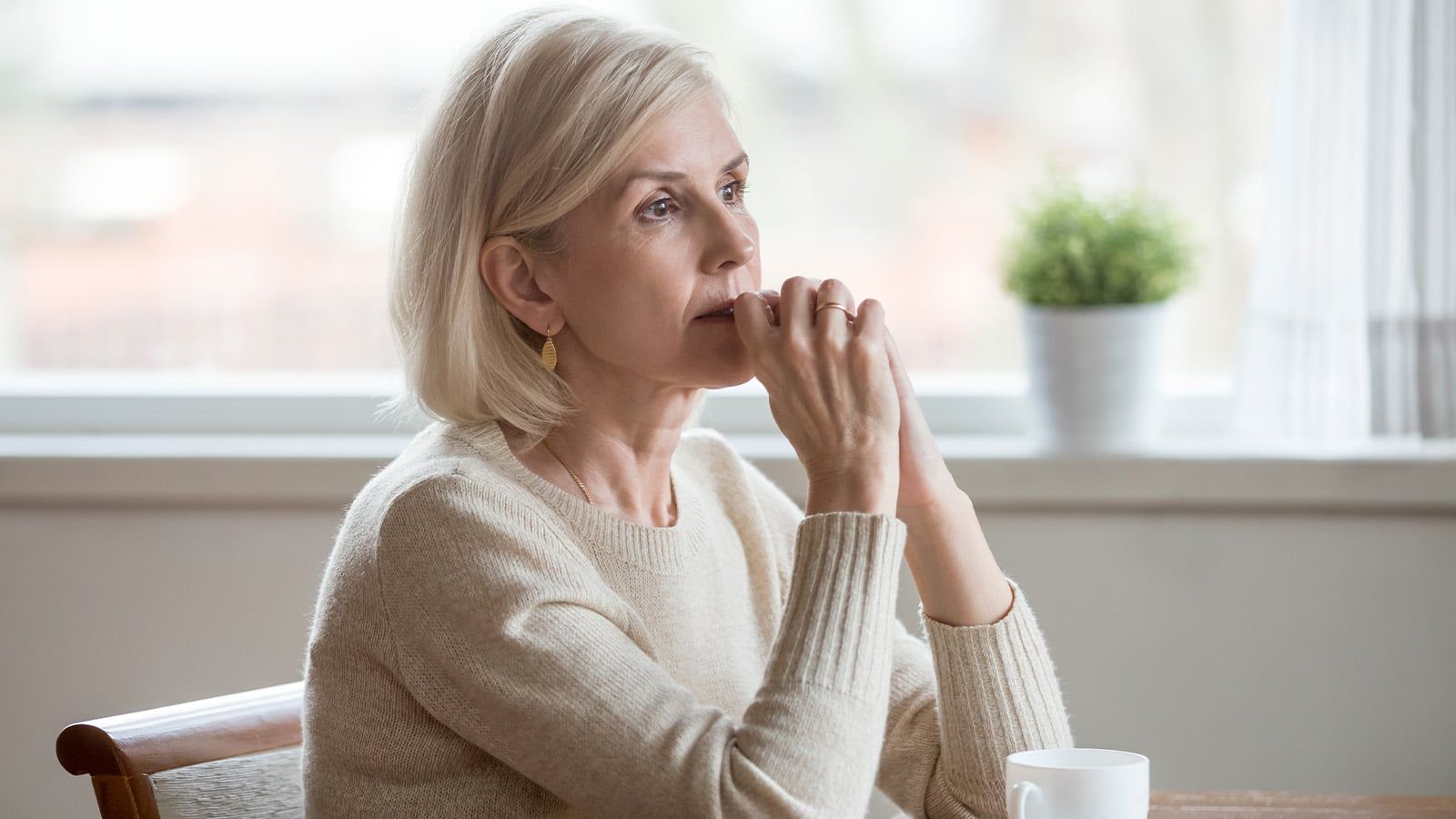 A woman with short blonde hair sits at a table indoors, resting her chin on her hands and looking thoughtfully out of the window. A white cup and a potted plant are visible in the background.