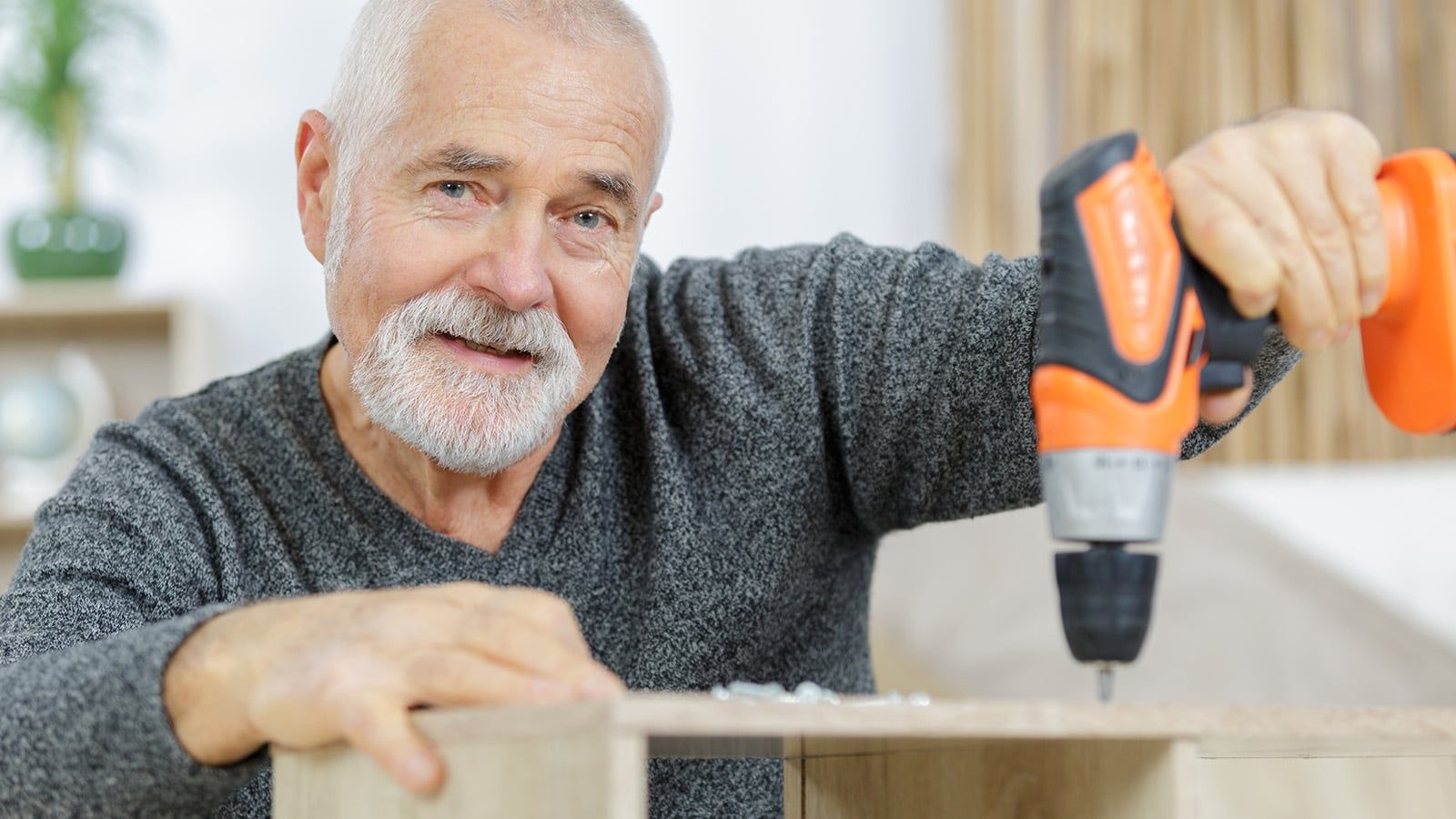 An older man with gray hair and a beard smiles while using an orange cordless drill to assemble a wooden piece of furniture indoors.