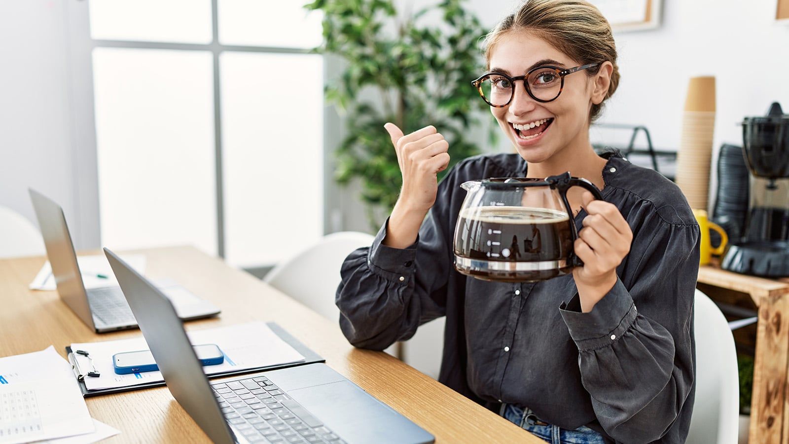A smiling person wearing glasses sits at a desk with laptops, holding a large coffee pot in one hand and giving a thumbs up with the other. The workspace has plants and coffee supplies in the background.
