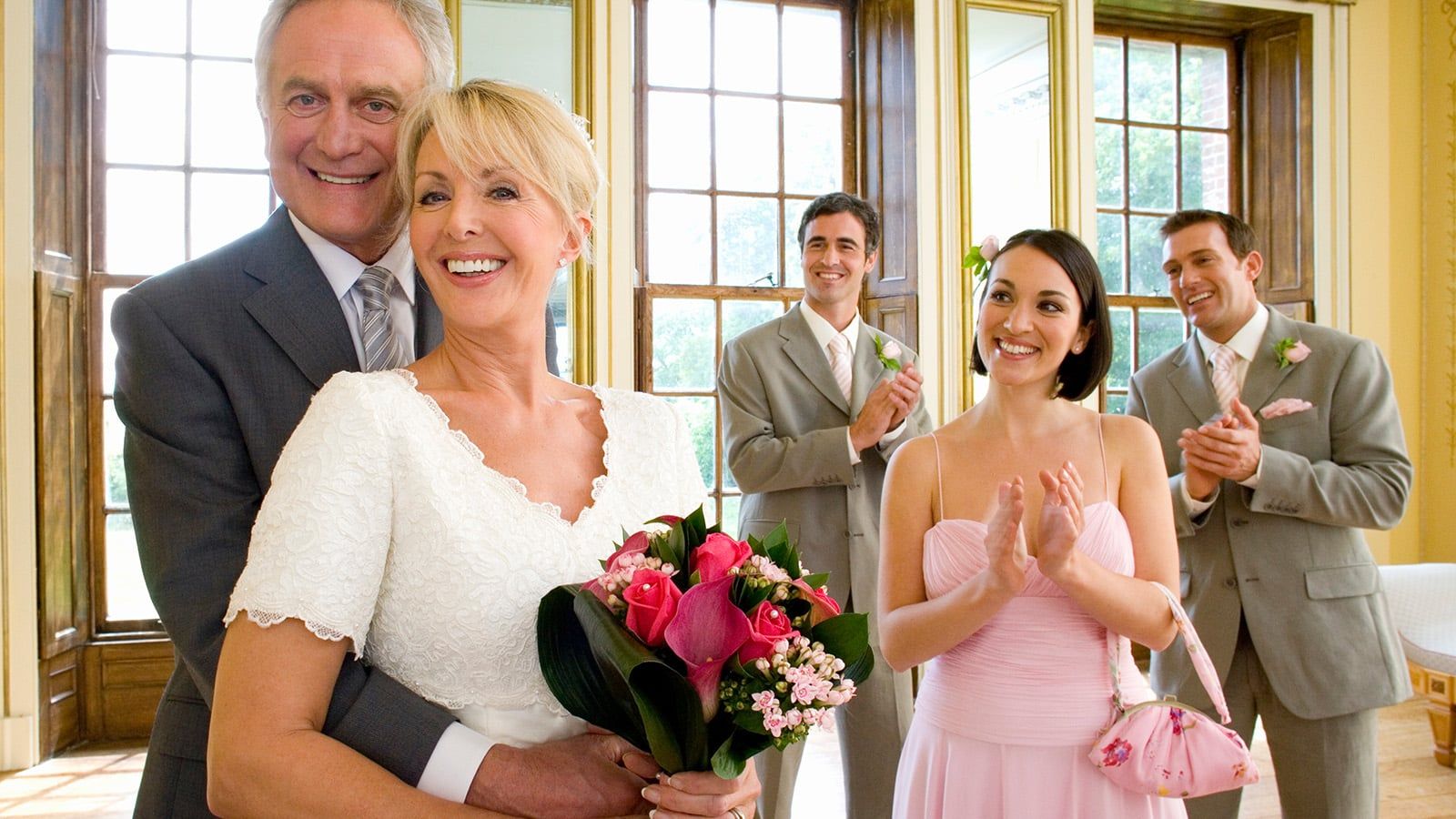 A smiling bride and groom embrace in a bright, elegant room, while three well-dressed guests—perhaps celebrating someone marrying into a family as second wives—stand behind them, clapping and sharing in the joyful moment.