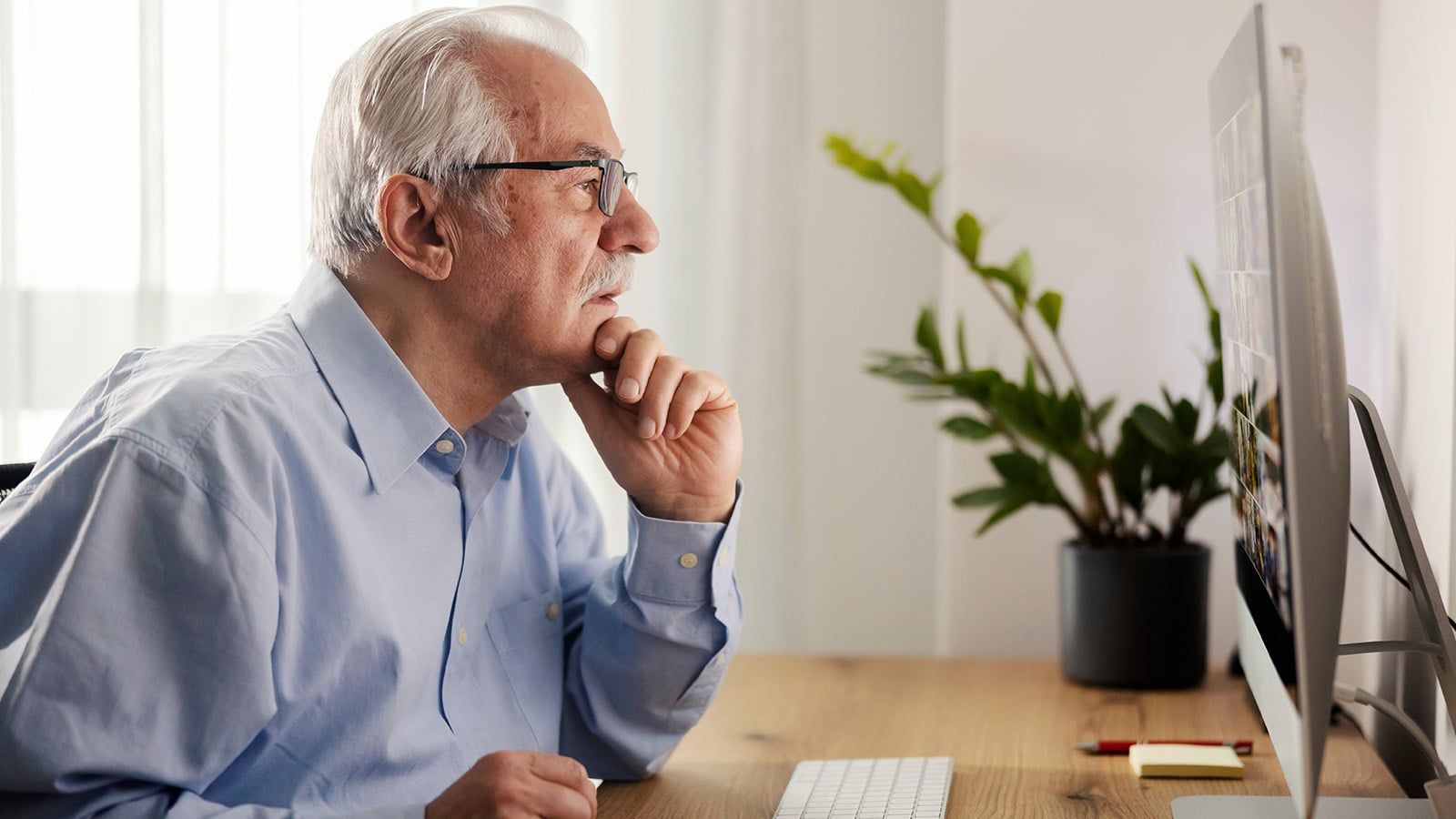 An older boomer dad with gray hair and glasses sits at a desk, looking intently at a computer monitor—perhaps scrolling through Facebook. He rests his chin on his hand, beside a potted plant and keyboard.