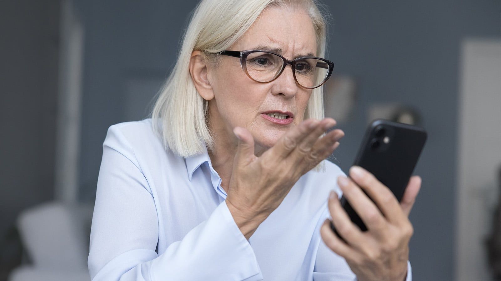 An older woman with gray hair and glasses looks frustrated while holding a smartphone, perhaps confused by modern social rules that seem second nature to Boomers or Gen X but not to her.