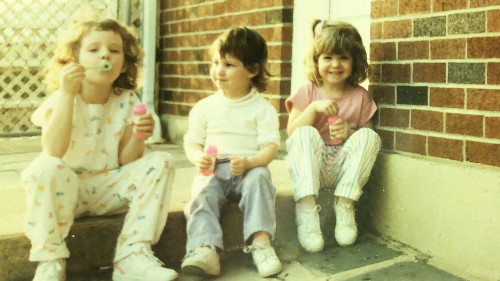 Three young children sit on outdoor steps blowing bubbles, smiling and holding bubble bottles. Dressed in casual clothes and sneakers, they capture the carefree spirit of a self-sufficient generation against a brick wall and fence backdrop.