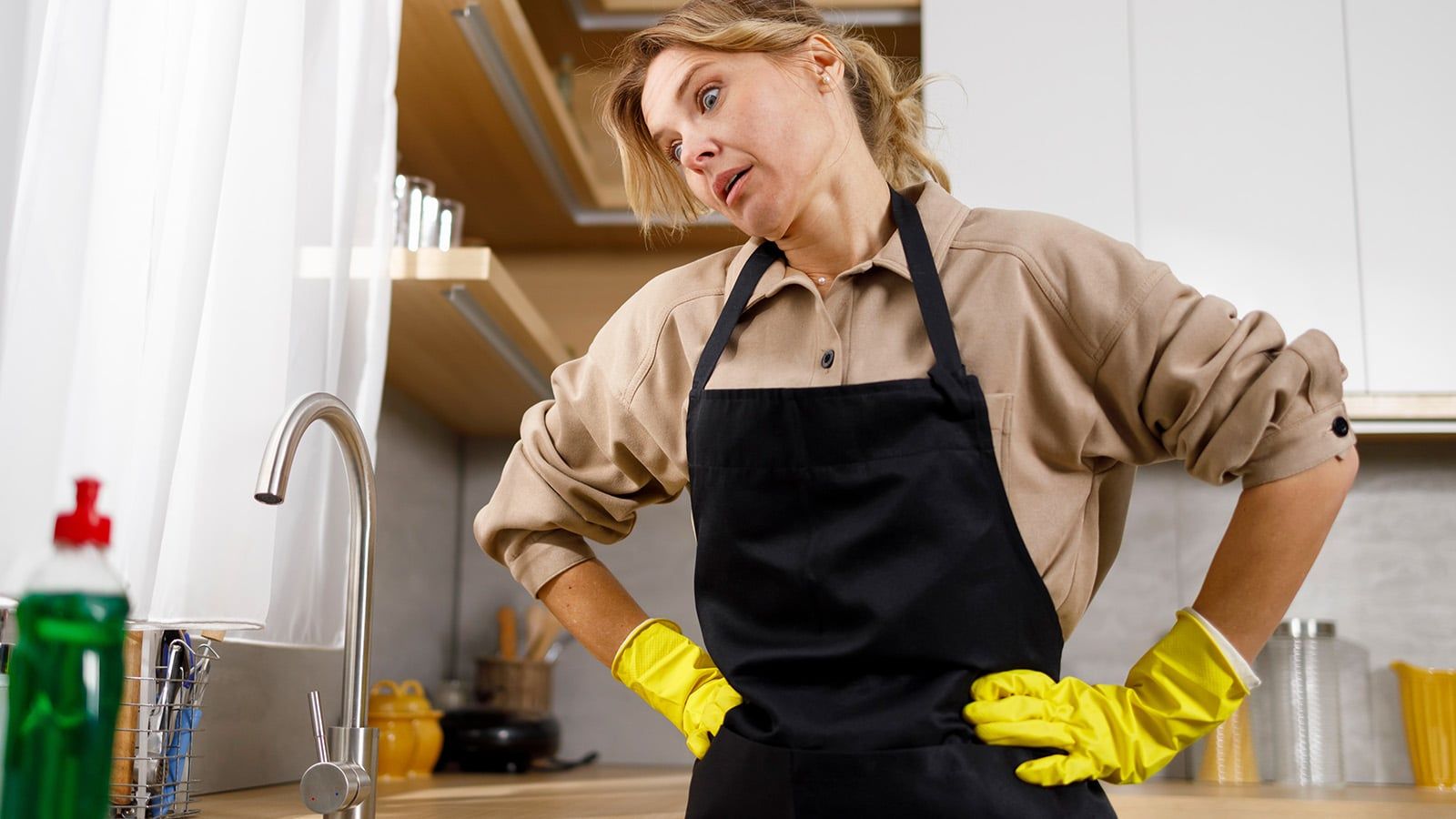 A woman in an apron and yellow gloves stands in a kitchen, looking tired with her hands on her lower back beside a sink and dish soap. Sometimes, after years of life achievements, she embraces the freedom at 64 by letting the dishes sit just a little longer.
