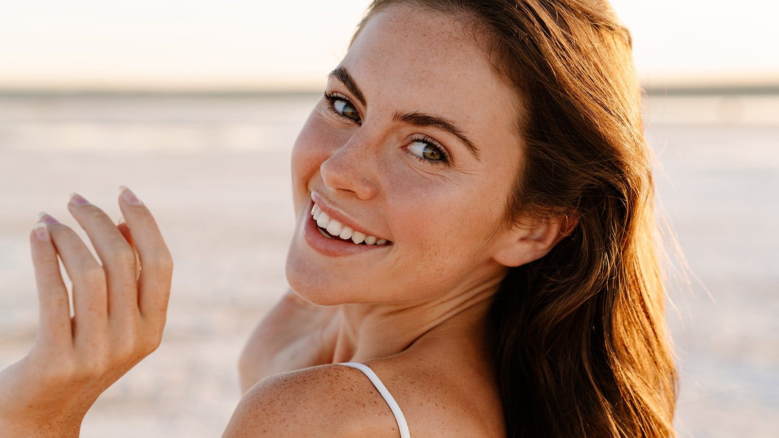 A woman with long brown hair smiles at the camera outdoors, sunlight illuminating her face. She is wearing a white top and standing on a sandy background, radiating the warmth and openness that are signs of trust and making her seem like a trusted person.
