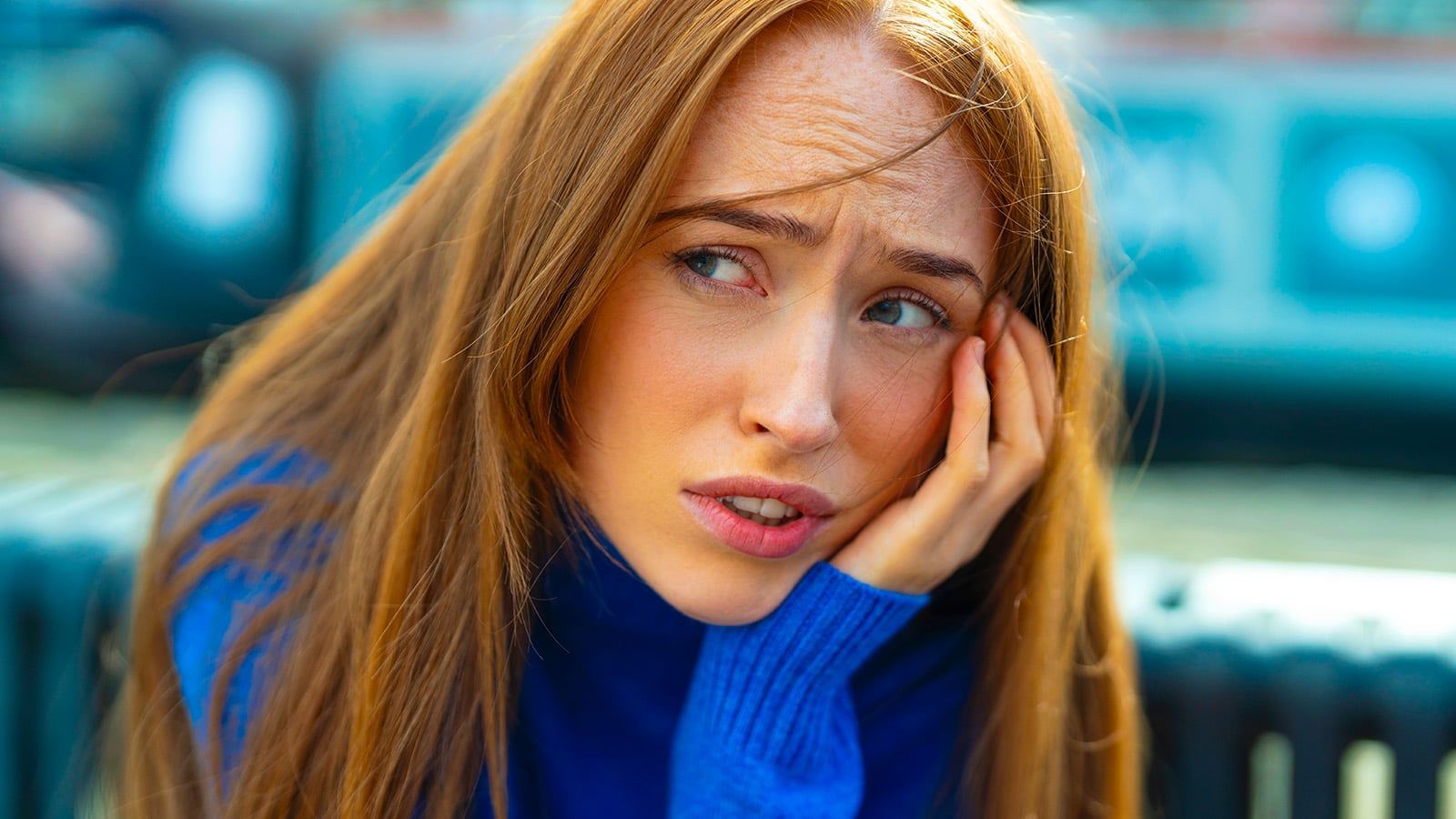 A woman with long red hair wearing a blue sweater looks worried, resting her face on her hand. The background is blurred, capturing the quiet weight eldest daughters often feel as they navigate complex family roles.