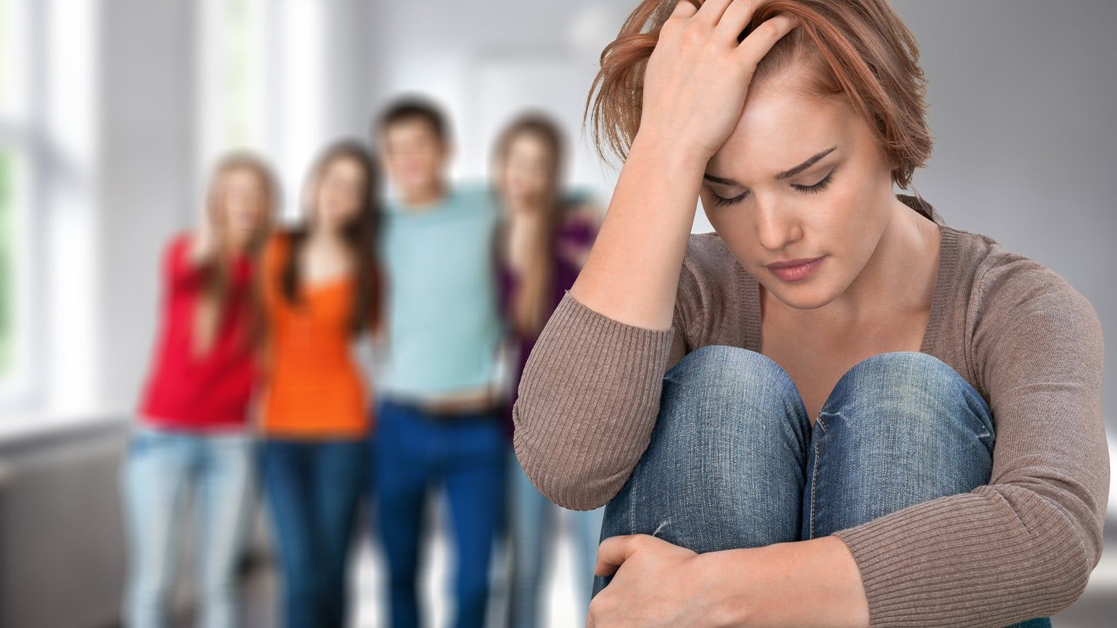 A young woman with rare traits sits alone, looking upset with her head resting on her hand, while a group of four people stands together in the blurred background.