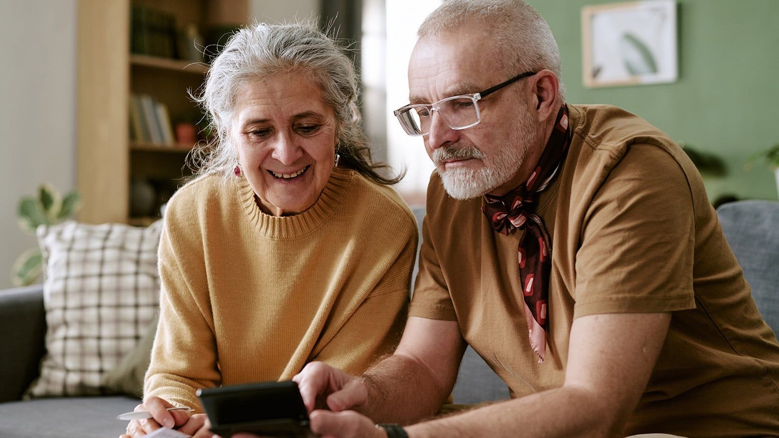 An older woman and man sit together on a couch, smiling at a smartphone. Despite past financial anxiety, they now enjoy the warmth of their cozy living room. The woman wears a tan sweater; the man wears glasses, a brown shirt, and a scarf.