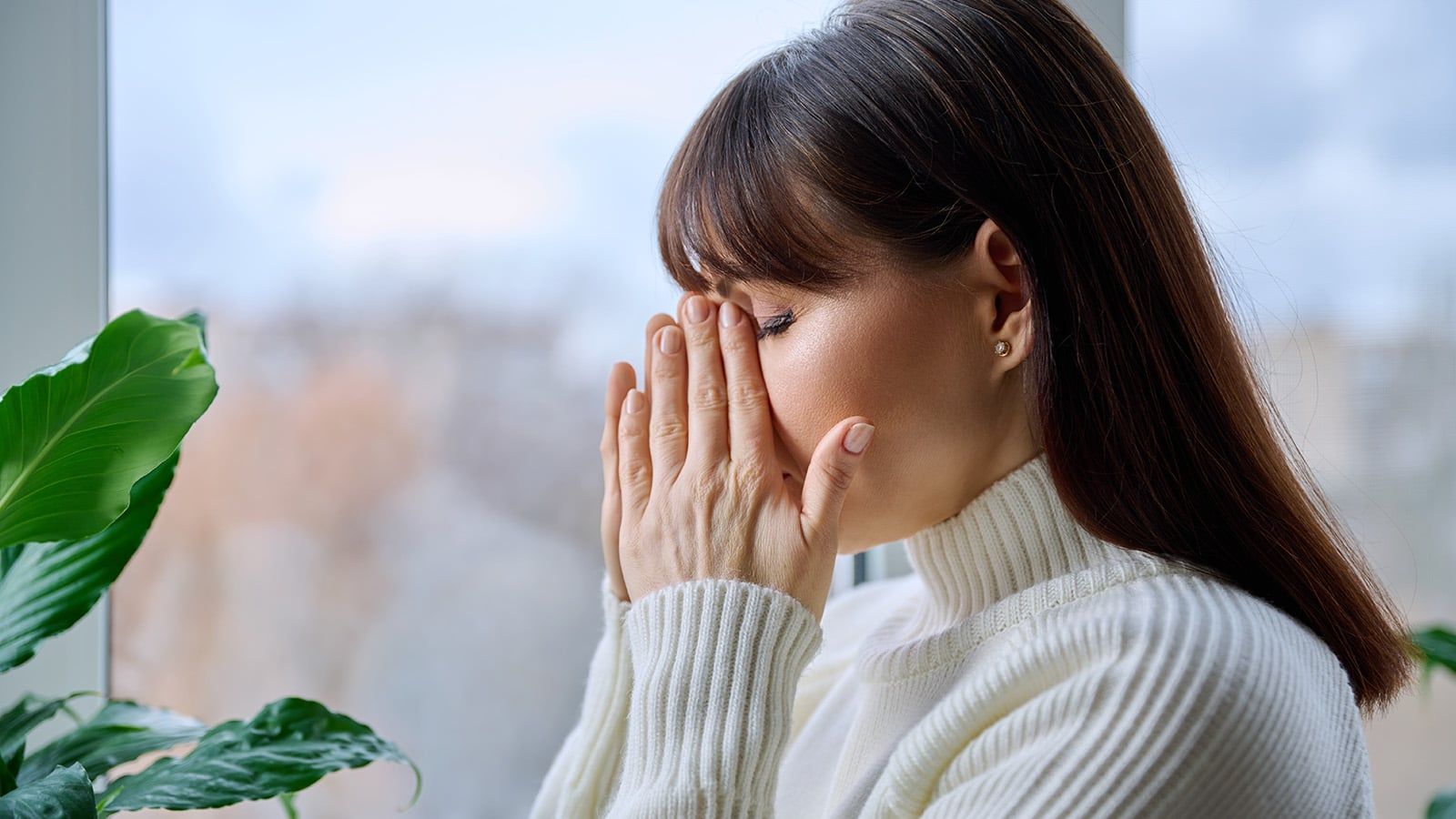 A woman in a white turtleneck sits by a window with her eyes closed and hands covering her nose and mouth, appearing stressed or upset—perhaps overwhelmed by the mental load of household management. A green houseplant is visible beside her.