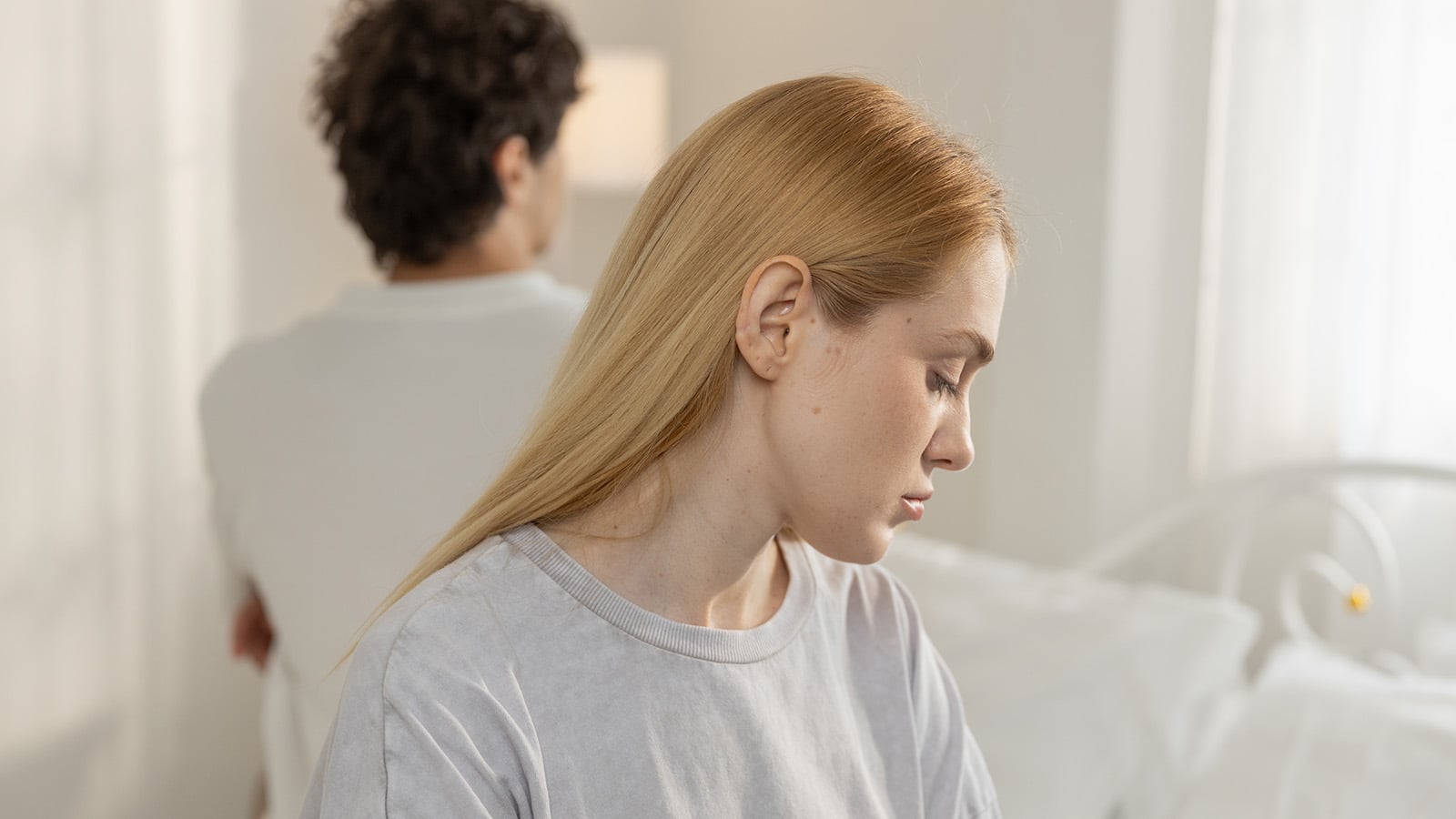 A woman with blonde hair looks down sadly while a man with dark curly hair sits behind her, facing away. Both appear upset in a bright bedroom, highlighting relationship challenges and moments of marriage loneliness.