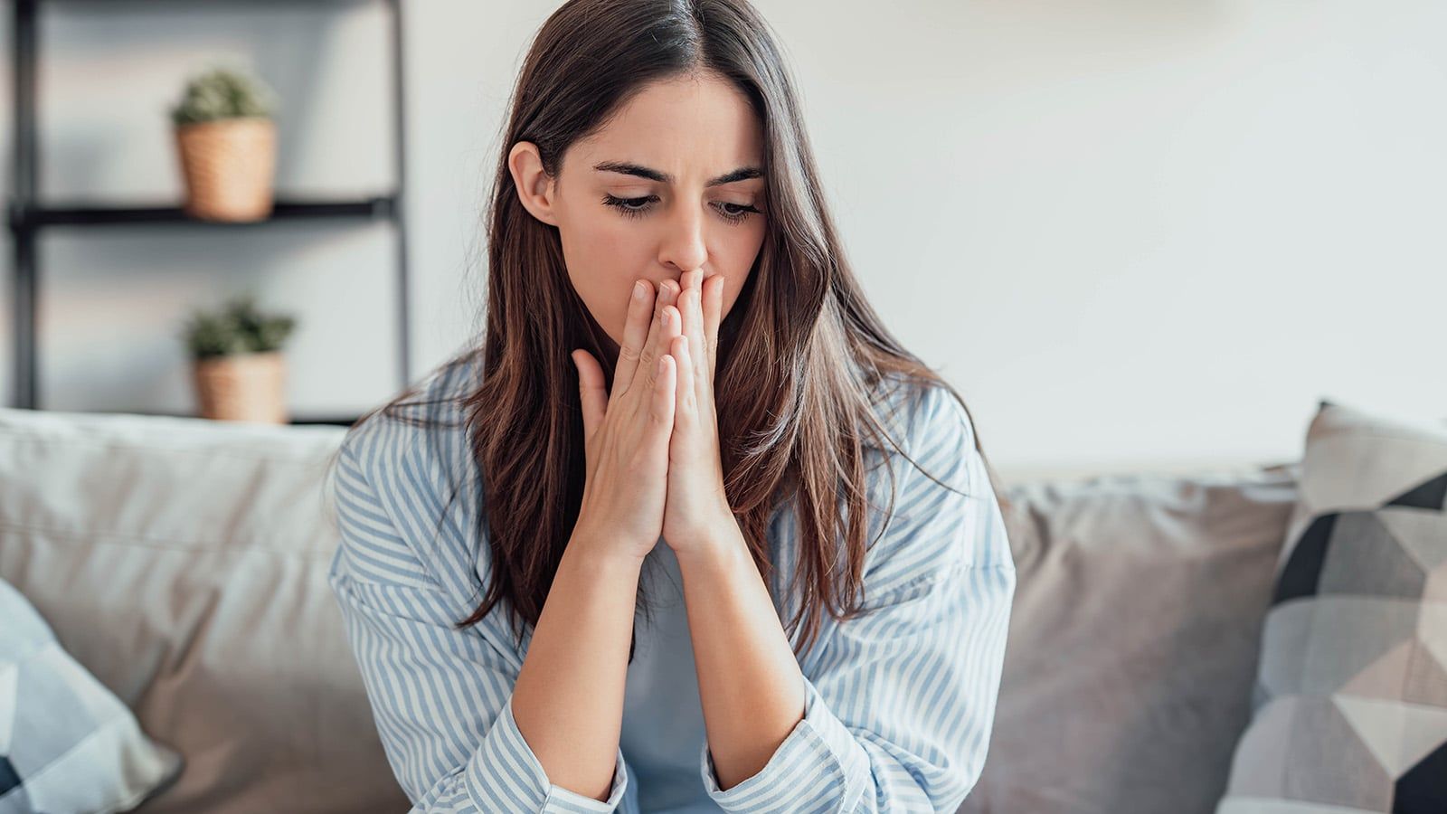 A young woman sits on a couch indoors, pressing her hands to her mouth and looking down with a worried or thoughtful expression, perhaps contemplating the emotional risk that often comes with maintaining high standards in relationships. Potted plants and shelves are visible in the background.